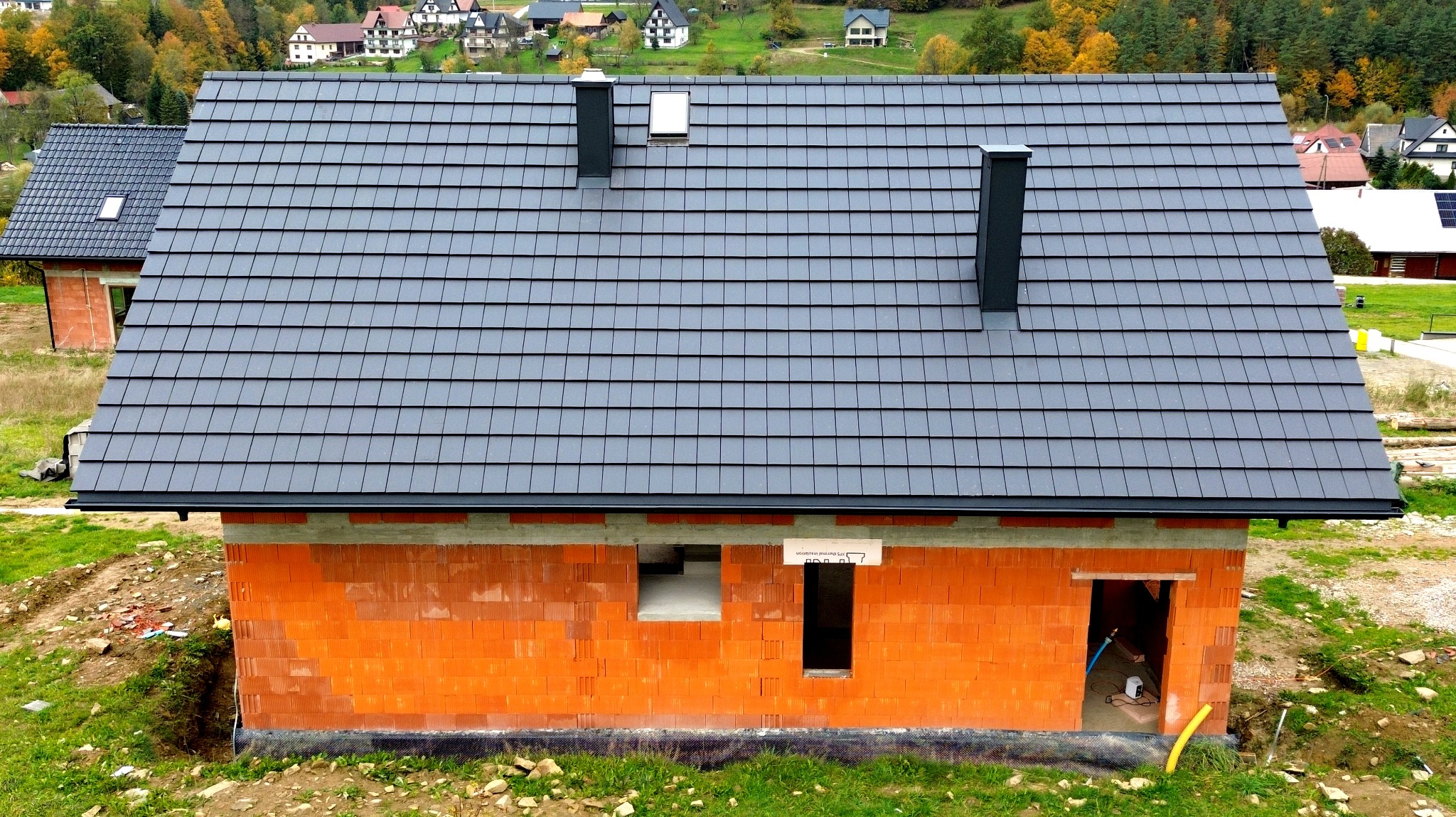 Dom w trakcie budowy z pomarańczowych pustaków, z szarą dachówką, czarnymi kominami i oknem dachowym, widoczne elementy instalacji wewnątrz.