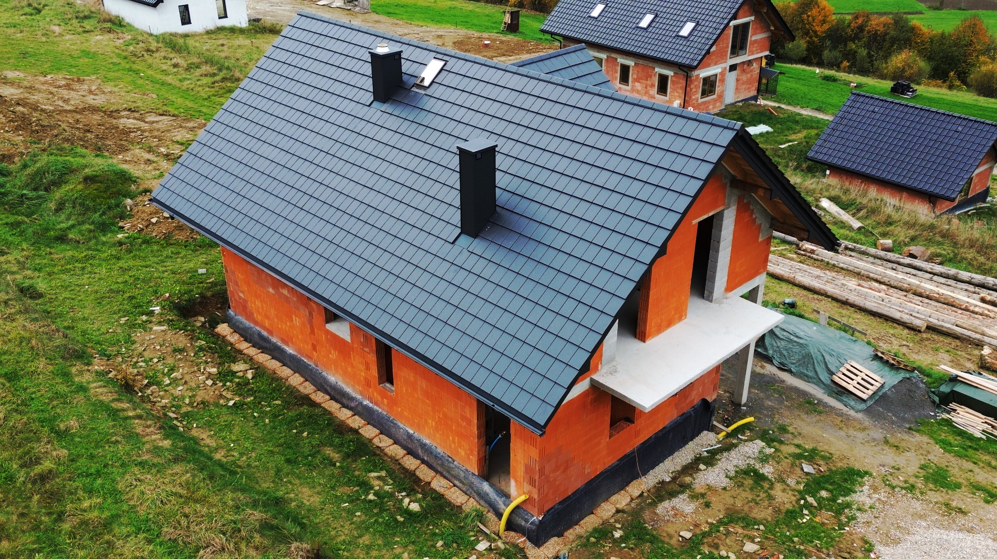 Niedokończony, murowany dom jednorodzinny z szarą dachówką i dwoma kominami, widok z góry. W tle inne budynki w różnym stadium zaawansowania budowy oraz teren zielony.