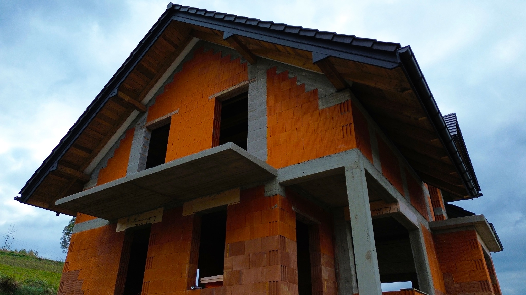 Surowy stan domu z czerwonej cegły, widoczne betonowe nadproża okienne i zadaszenie z ciemnej dachówki na tle pochmurnego nieba.