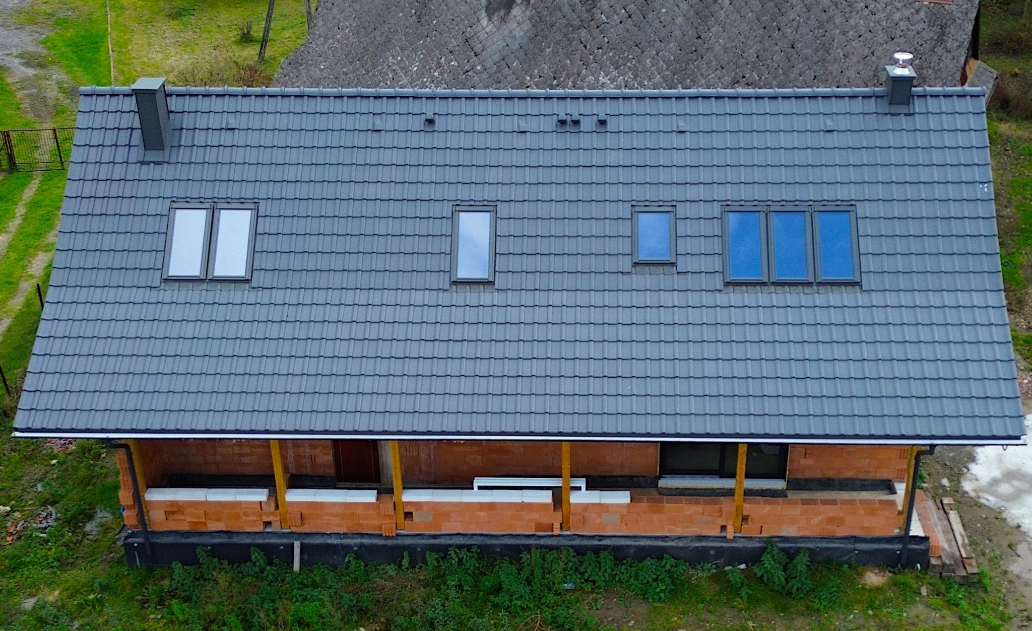 Widok z góry na dom w trakcie budowy, z szarym dachem z dachówki ceramicznej, czterema oknami dachowymi różnej wielkości i ceglanymi ścianami parteru z drewnianymi podporami.