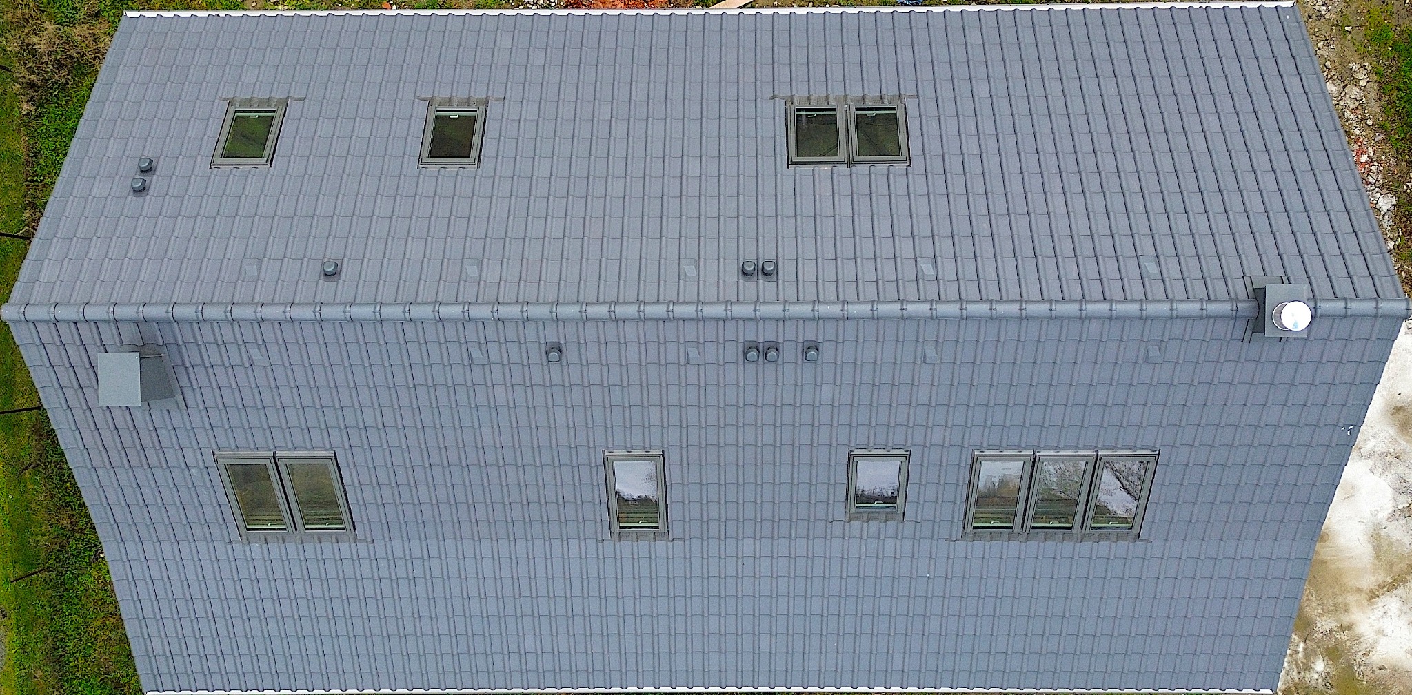 Widok z góry na skośny dach pokryty szarą dachówką ceramiczną z kilkoma oknami dachowymi, kominkami wentylacyjnymi i orynnowaniem