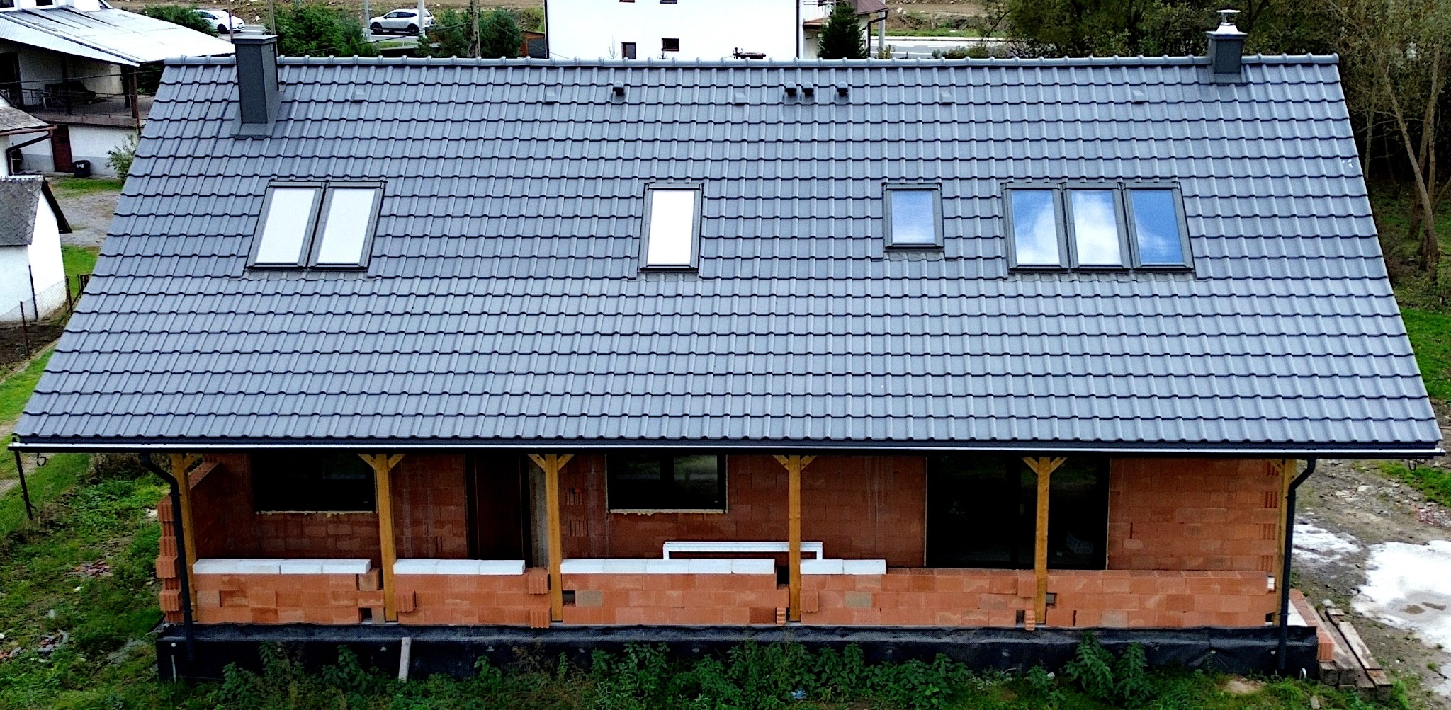 Dom w trakcie budowy z szarą dachówką, widoczne okna dachowe, ściany z czerwonej cegły i drewniane podpory werandy.
