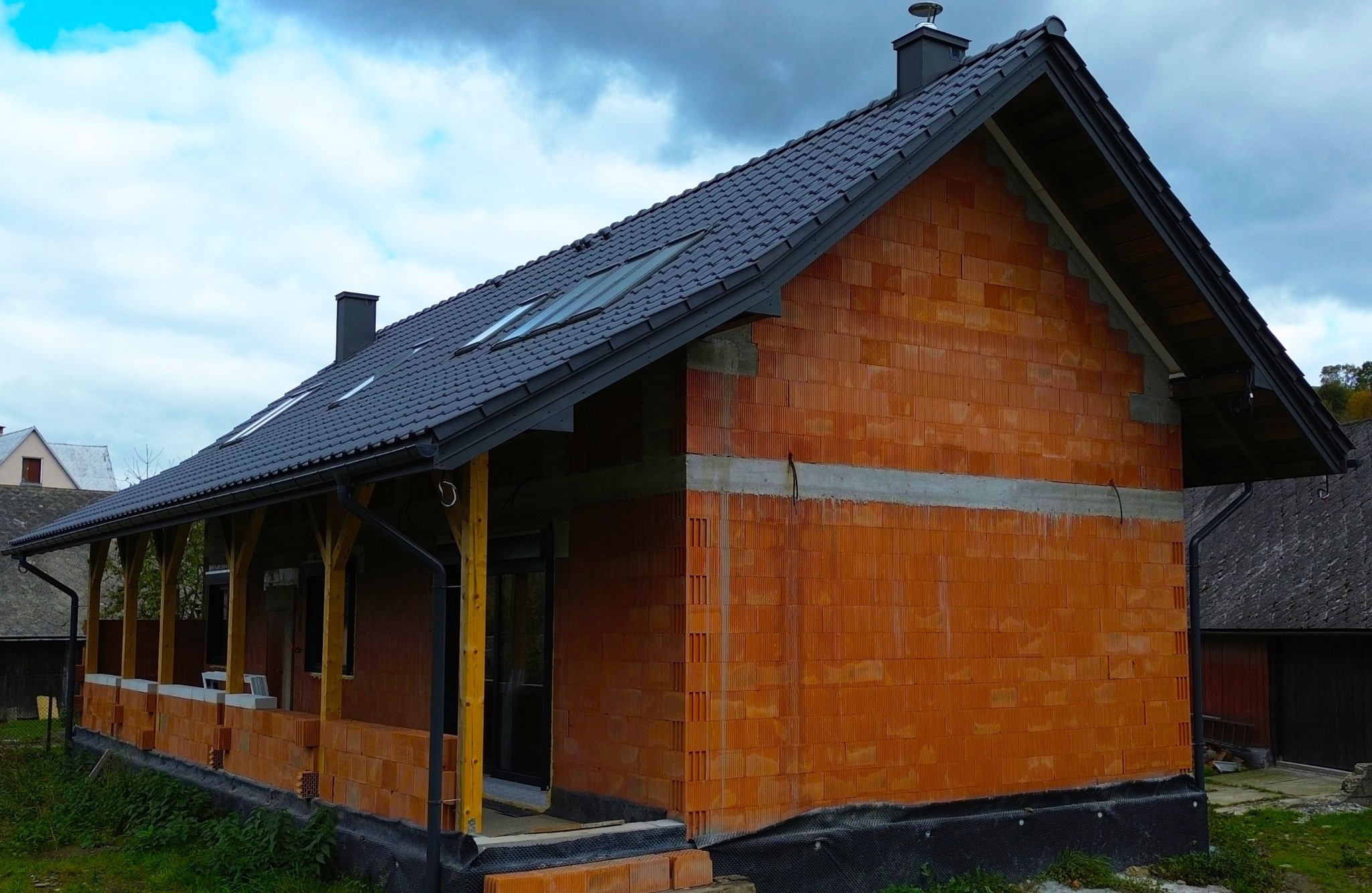 Surowy stan domu jednorodzinnego z czerwonej cegły ceramicznej, z czarną dachówką i drewnianą konstrukcją werandy, widok z zewnątrz na tle pochmurnego nieba.