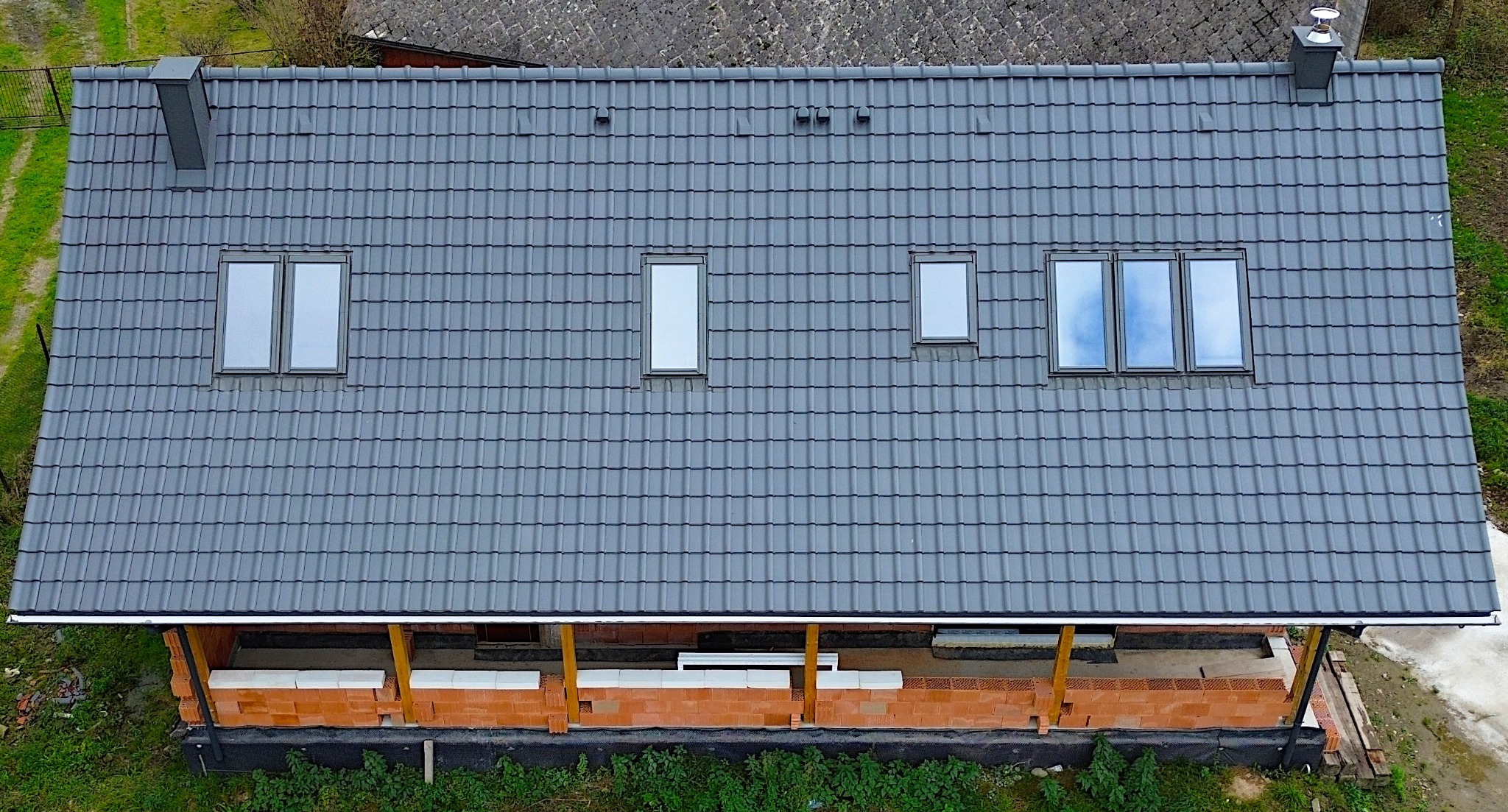 Widok z góry na dom w trakcie budowy z szarą dachówką, trzema oknami dachowymi różnej wielkości i kominem, ściany wzniesione z czerwonej cegły kratówki.