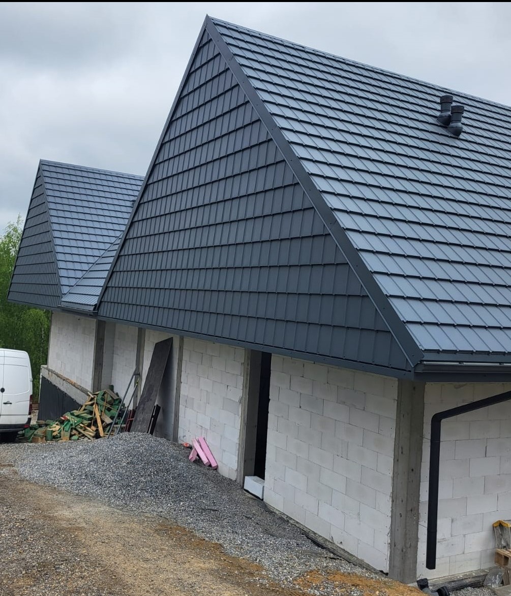 Surowy stan dwukondygnacyjnego budynku z szarego bloczka z ukończonym, szarym dachem z dachówki ceramicznej, widoczny stos materiałów budowlanych i zaparkowany biały van.