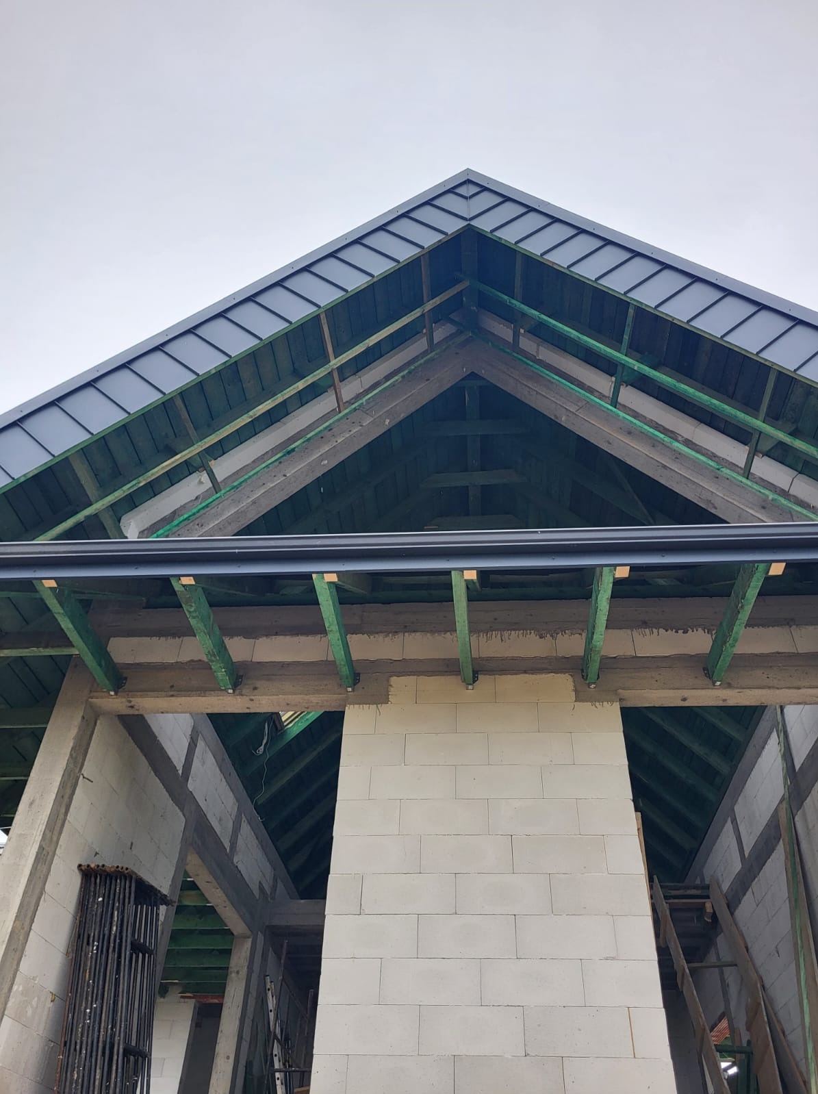 Wznoszenie dachu z szarej blachy na domu murowanym, widoczne krokwie i mur z bloczków betonowych w stanie surowym otwartym.