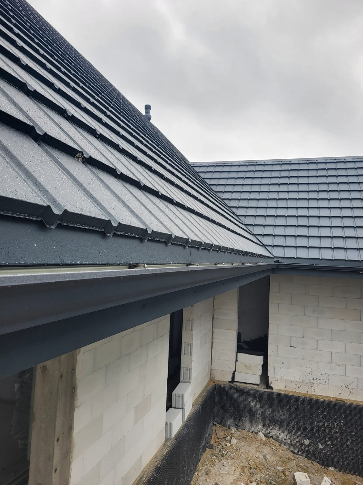 Szary dach z blachy trapezowej z widocznymi kroplami deszczu, orynnowanie w kolorze grafitowym, ściany z białego betonu komórkowego w trakcie budowy, widoczne otwory okienne i fundament...