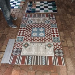 Sławomir filipiak - Układanie dekoracyjnych, wzorzystych płytek podłogowych na ceglanej posadzce, widoczne fragmenty ubrań osoby układającej i dodatkowe płytki w tle.