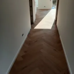 Korytarz z parkietem ułożonym w jodełkę, białe ściany z widocznymi gniazdkami elektrycznymi, wpadające światło dzienne.