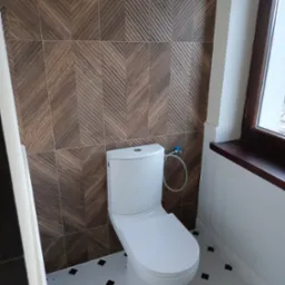 Nowoczesna łazienka z białą toaletą, brązowymi płytkami ściennymi ułożonymi we wzór jodełki i biało-czarną podłogą w karo, oświetlona naturalnym światłem z okna z ciemną ramą.