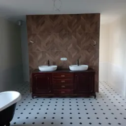 Łazienka w stylu retro z ciemną drewnianą szafką pod umywalki, białe umywalki nablatowe, brązowa ściana z płytek ułożonych w jodełkę, biało-czarna podłoga w karo, widoczna wanna na nóżkach...