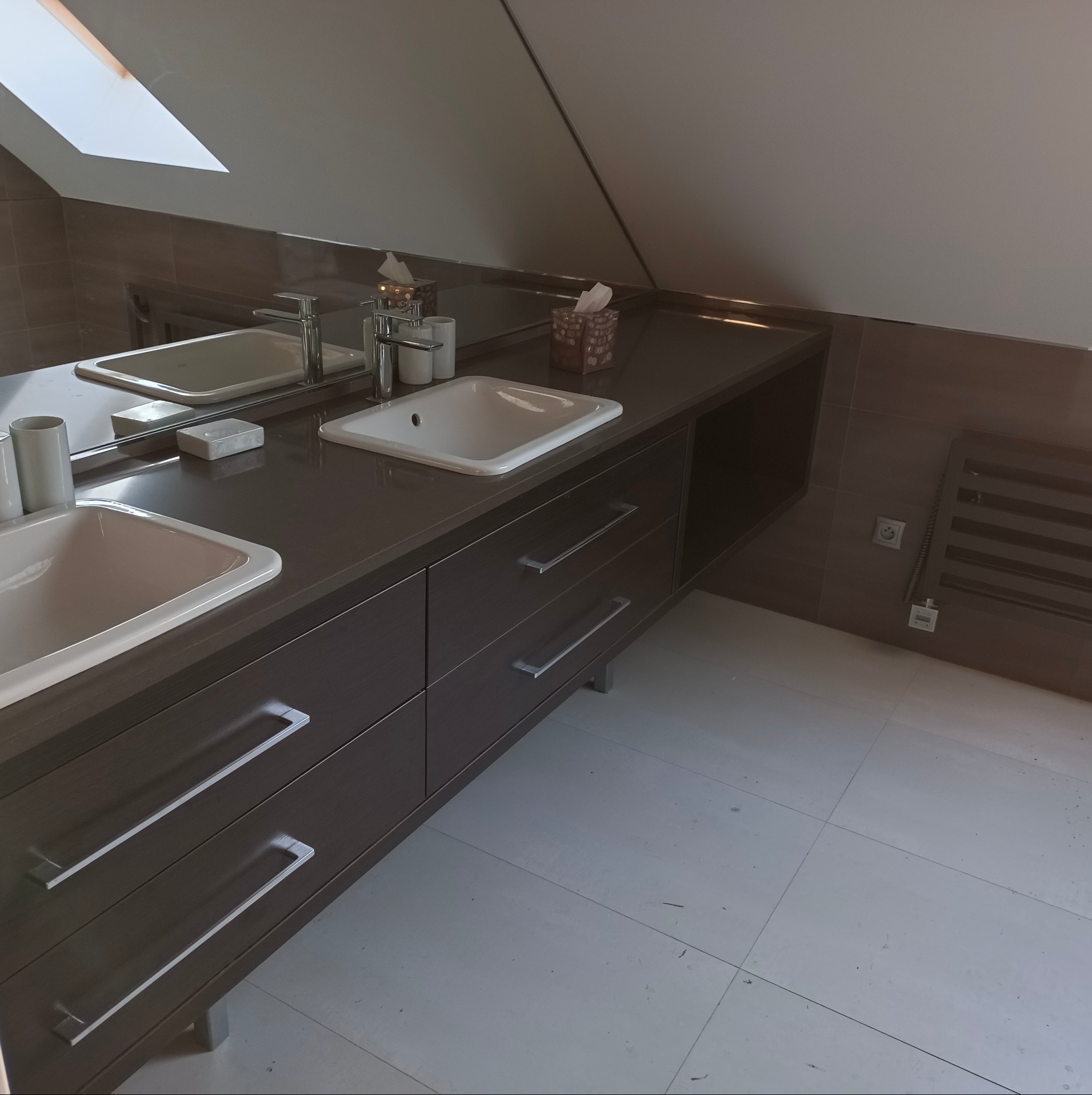 Nowoczesna łazienka na poddaszu z dwoma umywalkami nablatowymi, ciemnymi szafkami z długimi, srebrnymi uchwytami, dużym lustrem i grzejnikiem drabinkowym na ścianie wyłożonej brązowymi płytkami.