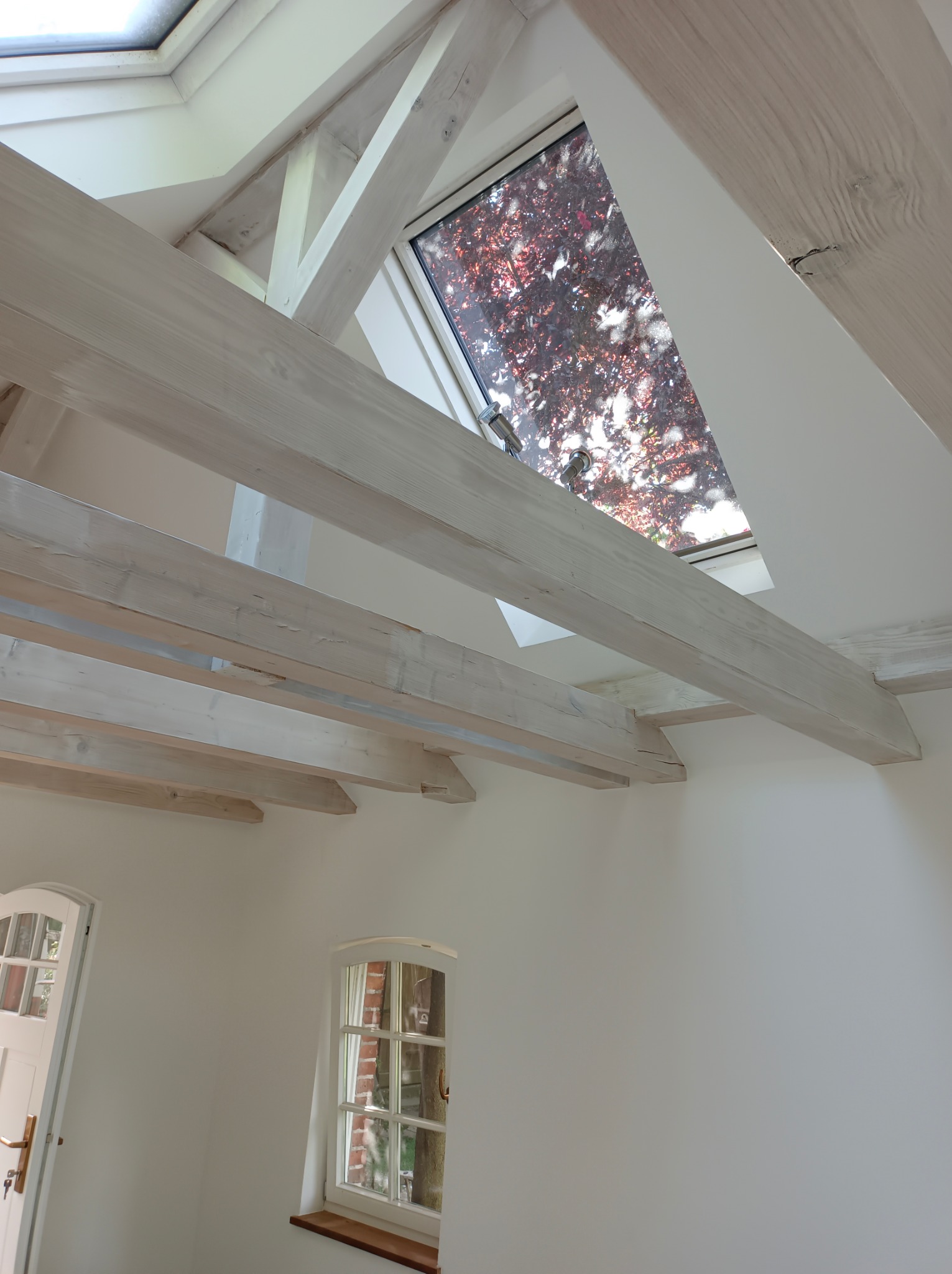 Białe, drewniane belki stropowe pod skośnym sufitem z oknem dachowym i małym oknem ściennym, widok z dołu.