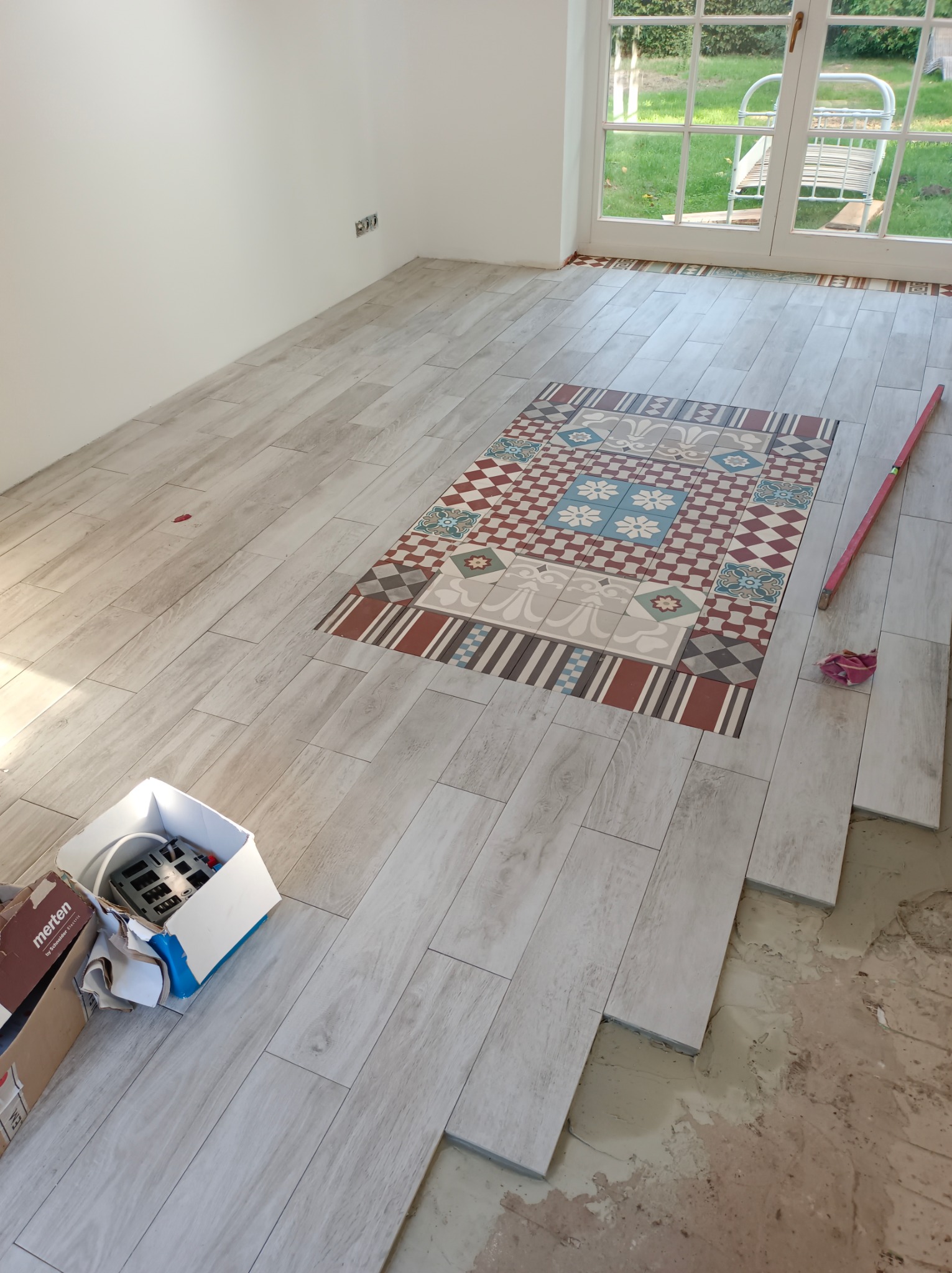 Układanie podłogi z płytek imitujących drewno z dekoracyjnym wzorem w centralnej części pomieszczenia, widoczny poziomica i pudełko z narzędziami.