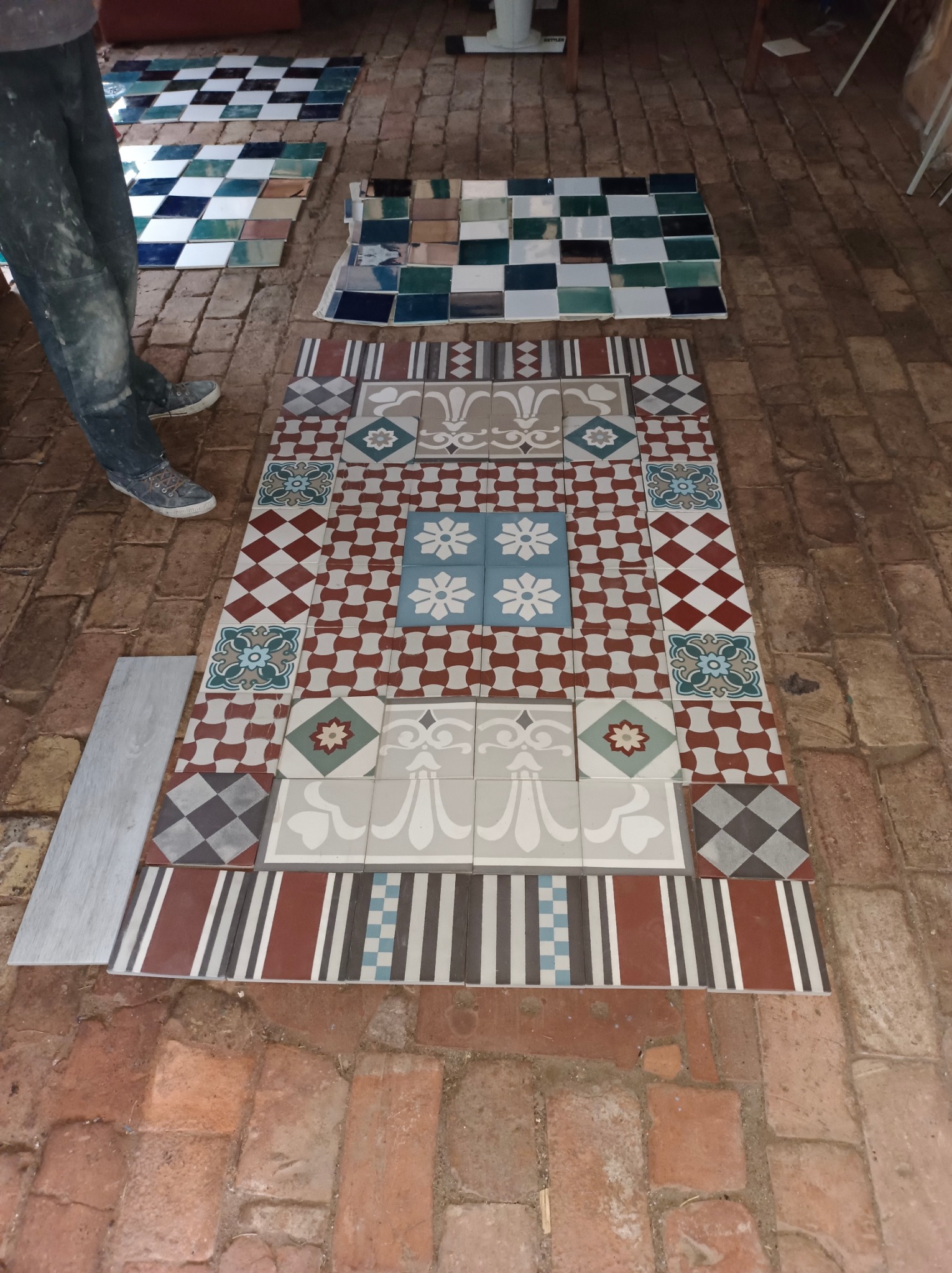 Układanie dekoracyjnych, wzorzystych płytek podłogowych na ceglanej posadzce, widoczne fragmenty ubrań osoby układającej i dodatkowe płytki w tle.