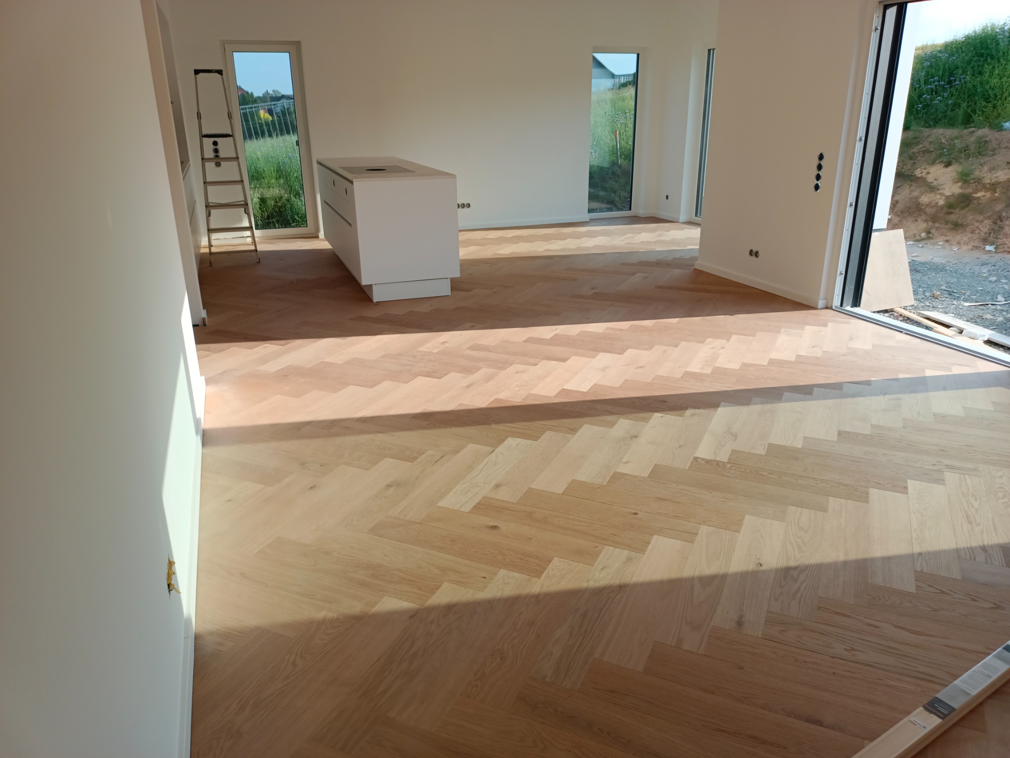 Jasny parkiet ułożony we wzór jodełki w przestronnym, jasnym wnętrzu z białymi ścianami i dużymi oknami; drabina oparta o ścianę w rogu, wyspa kuchenna w centrum, widok na zieleń za oknem.