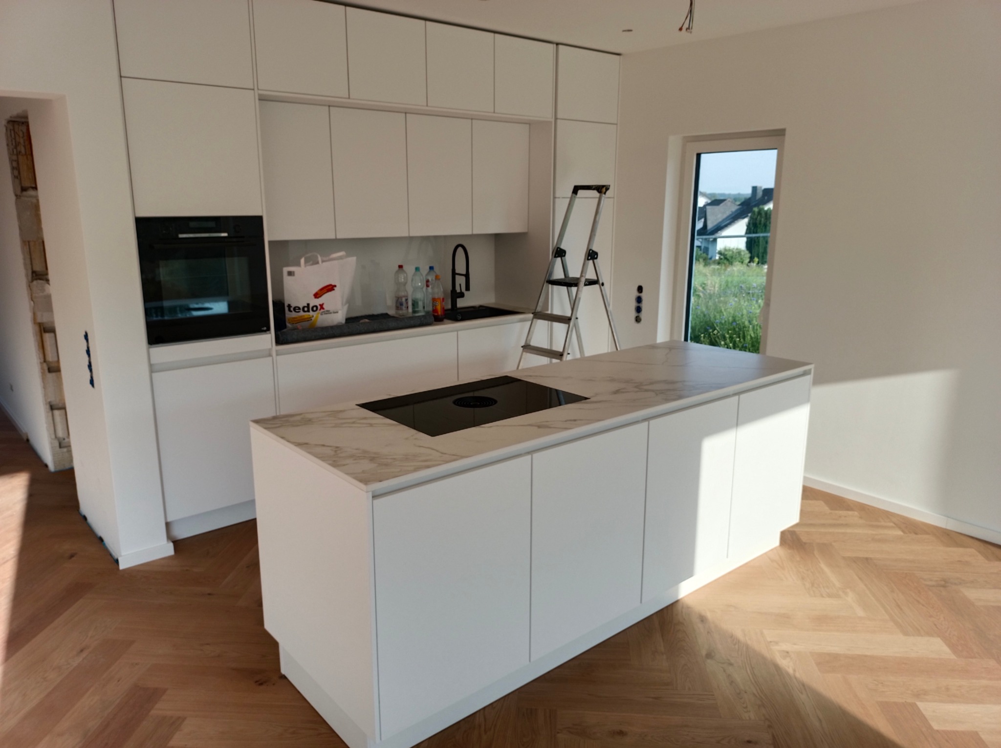 Nowoczesna, jasna kuchnia z białymi szafkami, wyspą z blatem imitującym marmur i parkietem w jodełkę, widoczna drabina malarska i torba zakupowa Tedox.