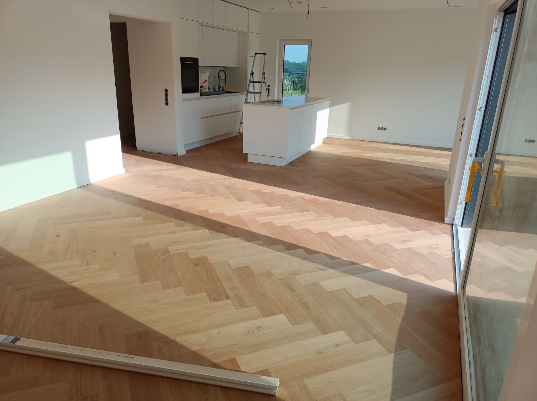 Jasny parkiet ułożony we wzór jodełki w nowym, przestronnym wnętrzu z białą kuchnią i wyspą kuchenną w tle, widoczne listwy przypodłogowe czekające na montaż.