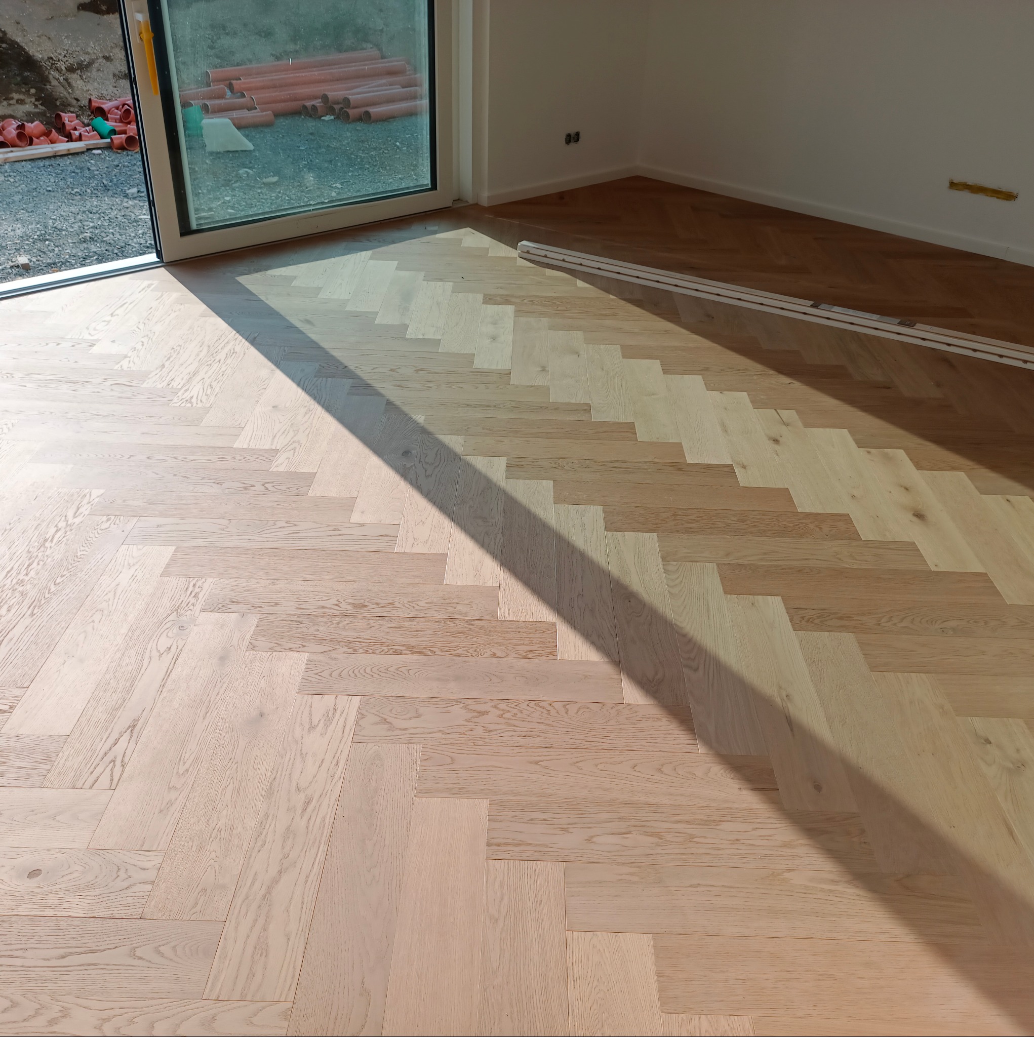 Jasna, drewniana podłoga ułożona we wzór jodełki, oświetlona silnym, ukośnym promieniem słonecznym w nowym, jasnym pomieszczeniu z widokiem na plac budowy przez okno.