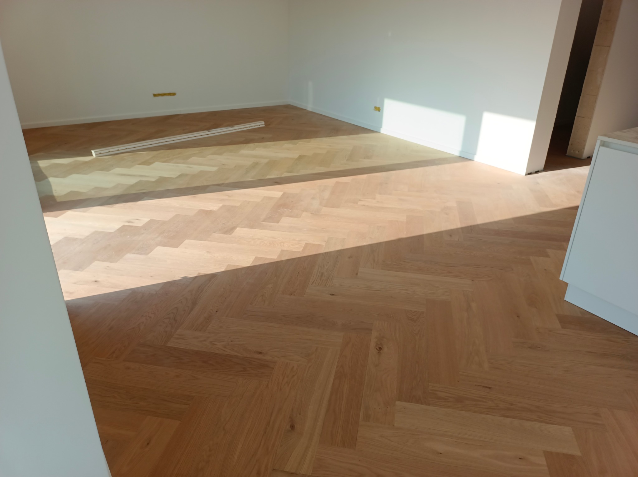 Parkiet ułożony w jodełkę w jasnym, pustym pomieszczeniu z białymi ścianami, listwy przypodłogowe w tle, fragment szafki w prawym rogu.