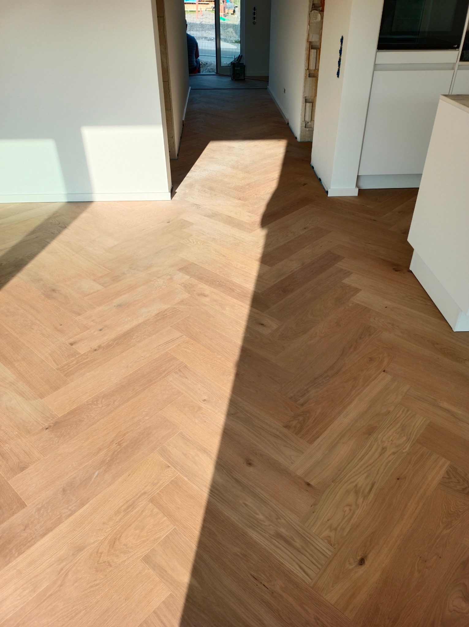 Jasna, drewniana podłoga ułożona w jodełkę, wpadające światło słoneczne, białe ściany w tle, widoczne fragmenty zabudowy kuchennej.