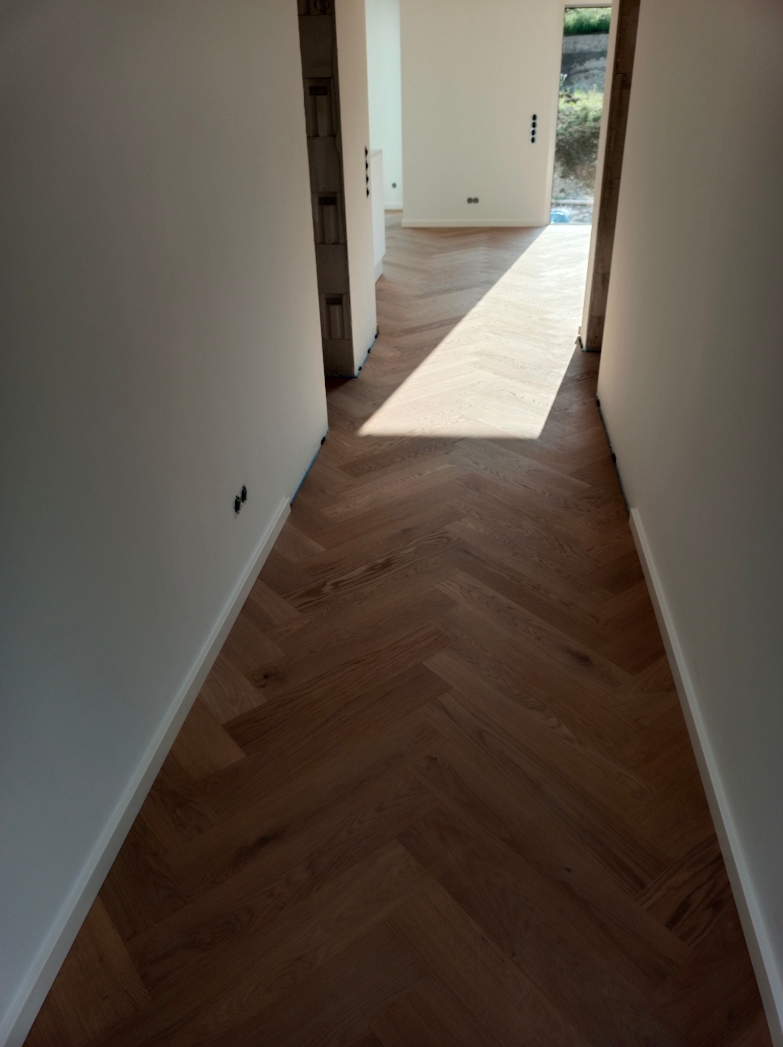 Korytarz z parkietem ułożonym w jodełkę, białe ściany z widocznymi gniazdkami elektrycznymi, wpadające światło dzienne.