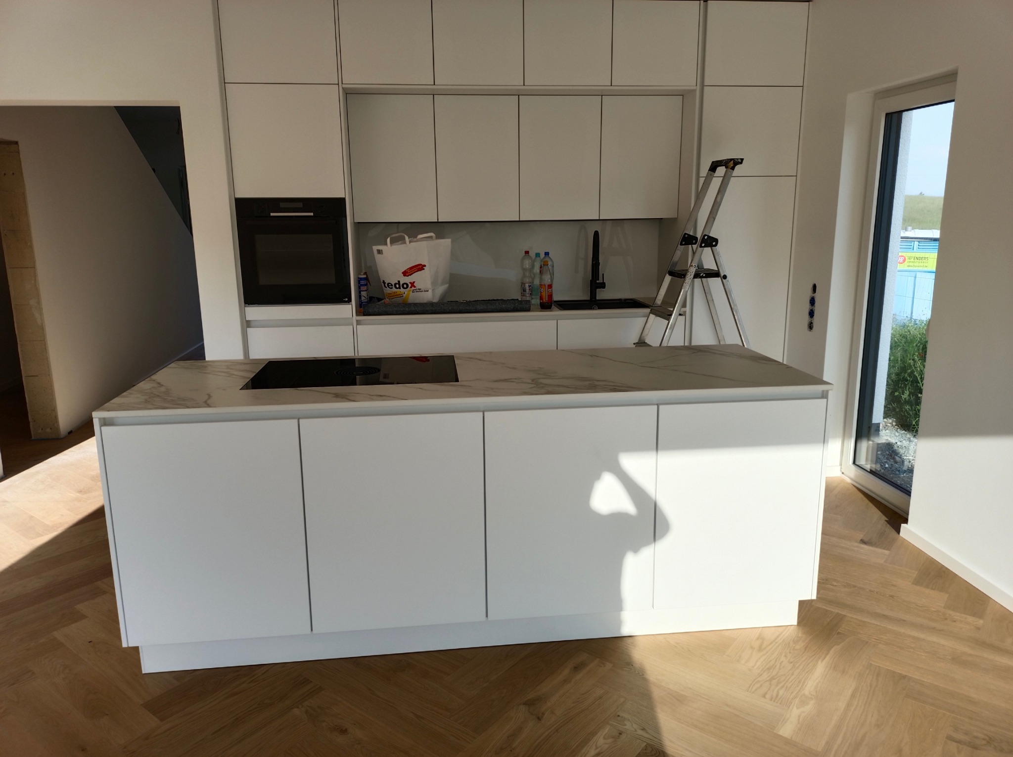 Nowoczesna, jasna kuchnia z białą zabudową meblową, marmurowym blatem wyspy, parkietem w jodełkę i widoczną drabiną malarską.