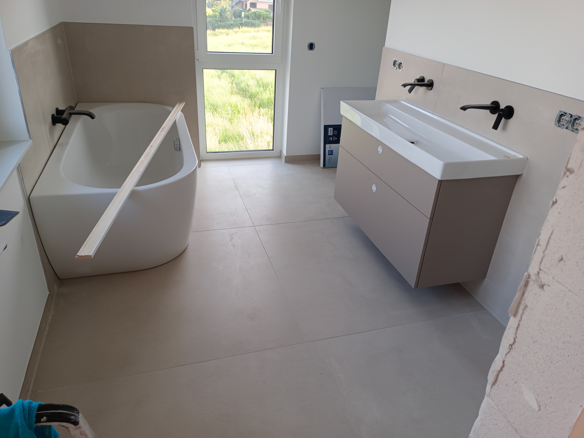 Nowoczesna łazienka w trakcie aranżacji: beżowe płytki, biała wanna z drewnianą listwą, szafka pod umywalkę z czarną armaturą, widoczne puszki elektryczne i fragment surowej ściany.