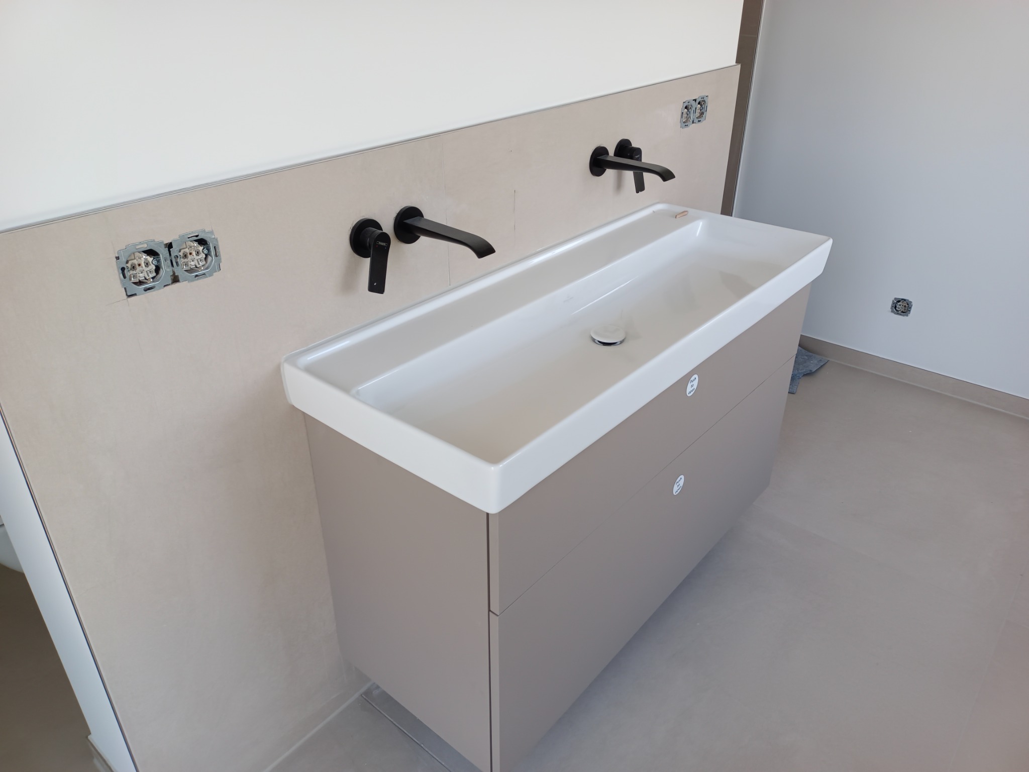 Nowoczesna umywalka z szafką w beżowym odcieniu, czarne baterie, niedokończone gniazdka elektryczne w tle, minimalistyczny design.