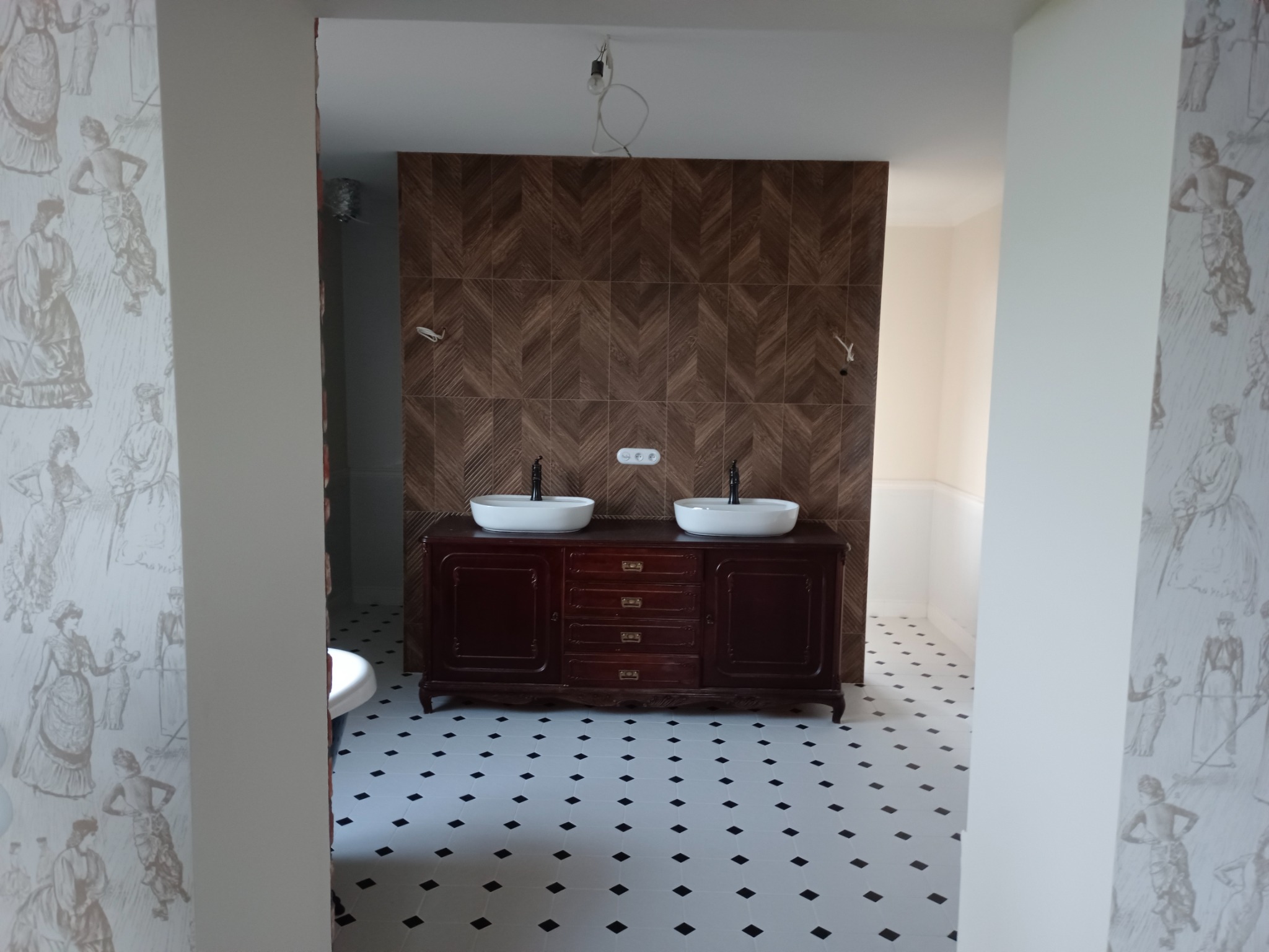 Łazienka w stylu retro z ciemną szafką pod umywalki, białe umywalki nablatowe, ściana wyłożona brązowymi płytkami w jodełkę, podłoga w biało-czarną szachownicę, tapeta w stylu vintage z postaciami.