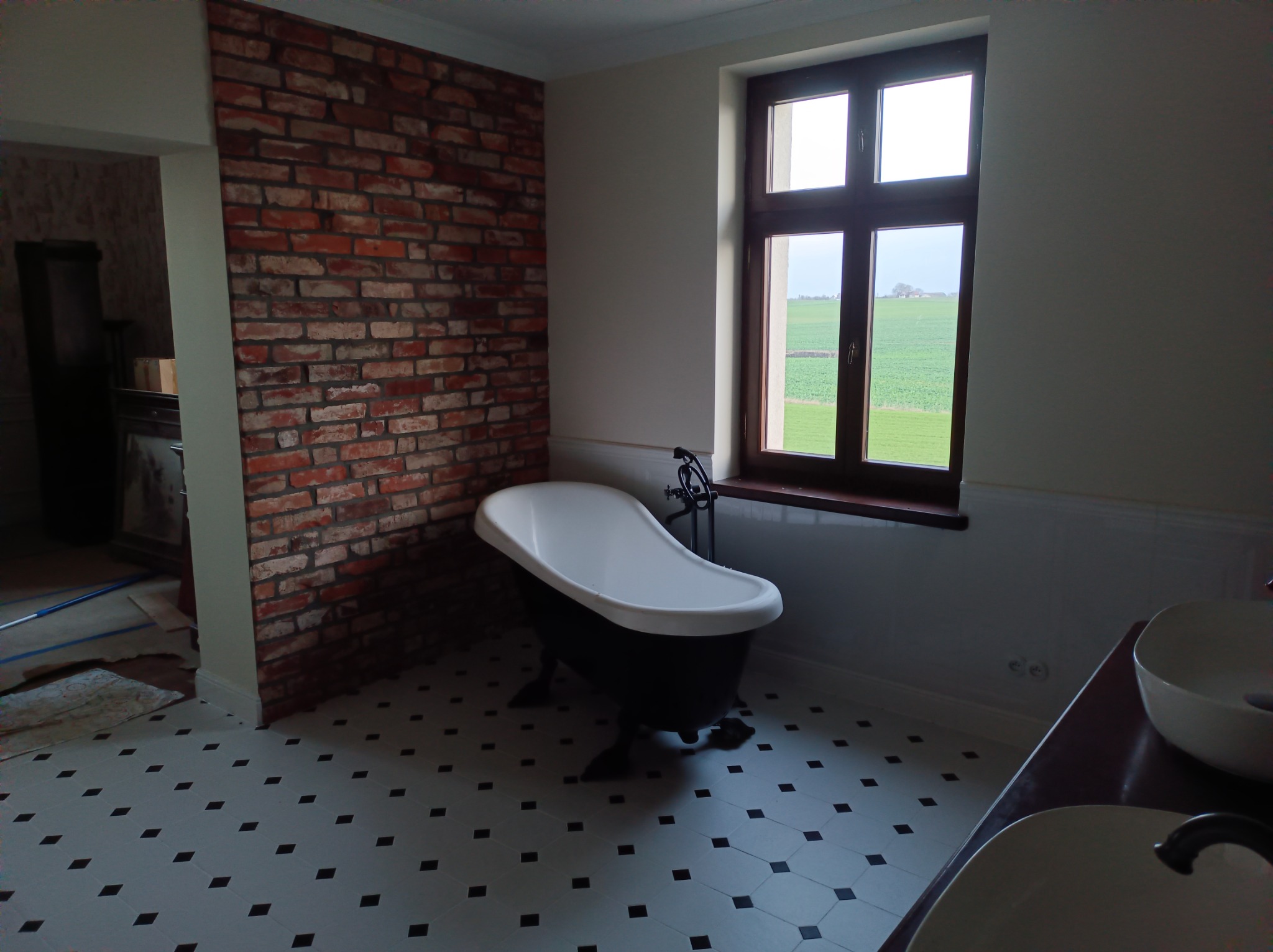 Retro łazienka z białą wanną na lwich nóżkach, ceglaną ścianą i oknem z widokiem na zielone pole za szybą.