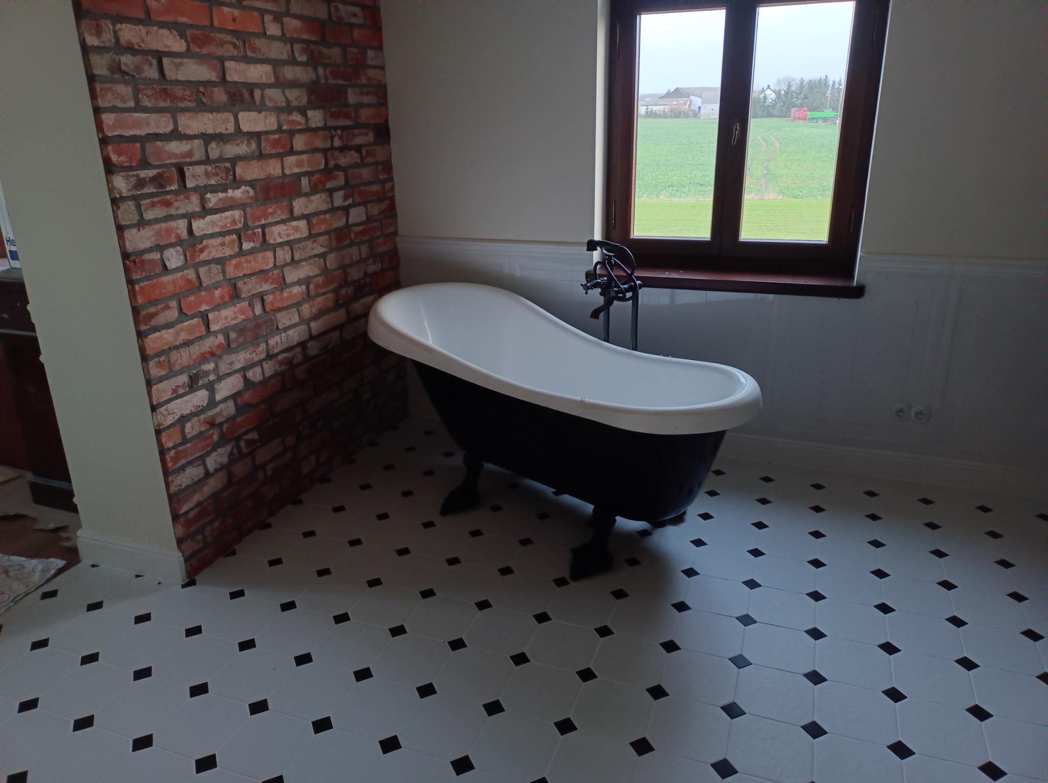 Wolnostojąca wanna na lwich nóżkach w łazience z ceglaną ścianą i widokiem na wiejski krajobraz za oknem.