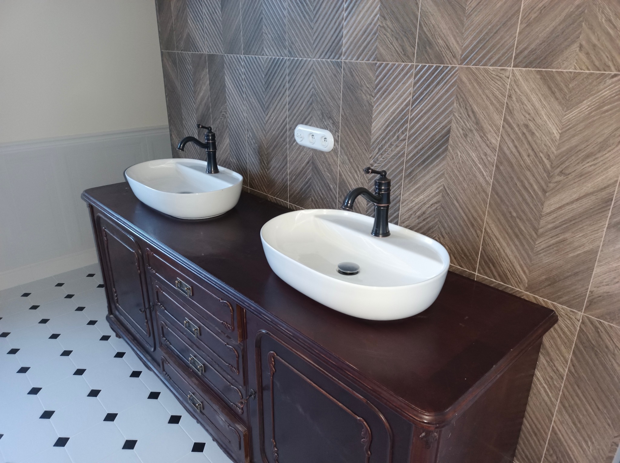 Dwie białe umywalki nablatowe z czarnymi bateriami na ciemnobrązowej, stylizowanej szafce z szufladami. W tle wzorzyste, szare płytki na ścianie i biało-czarna podłoga.