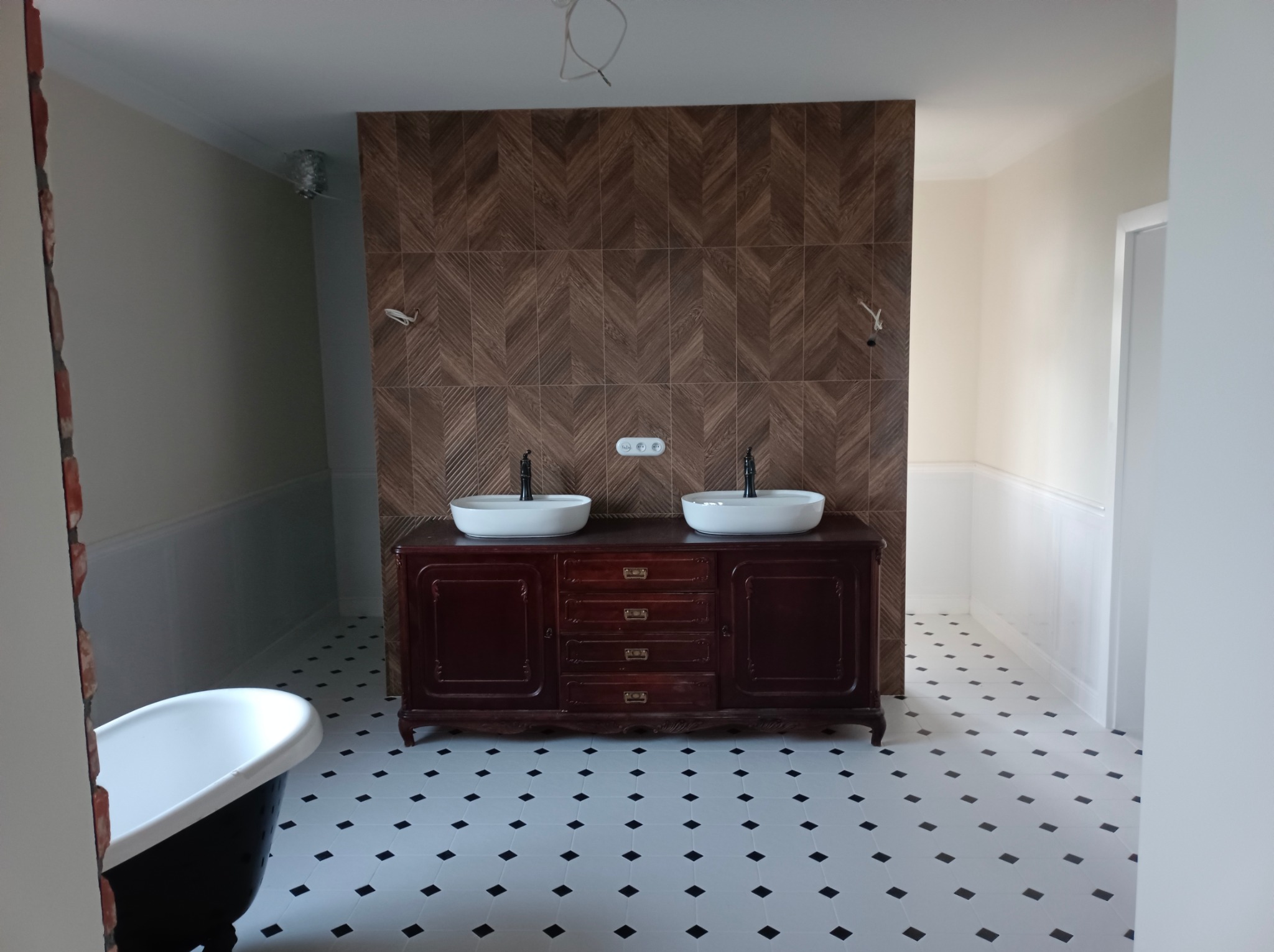 Łazienka w stylu retro z ciemną drewnianą szafką pod umywalki, białe umywalki nablatowe, brązowa ściana z płytek ułożonych w jodełkę, biało-czarna podłoga w karo, widoczna wanna na nóżkach...