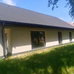 Nowoczesny parterowy dom z szarą dachówką, dużymi oknami i jasną elewacją, otoczony zielonym trawnikiem i drzewami w słoneczny dzień.