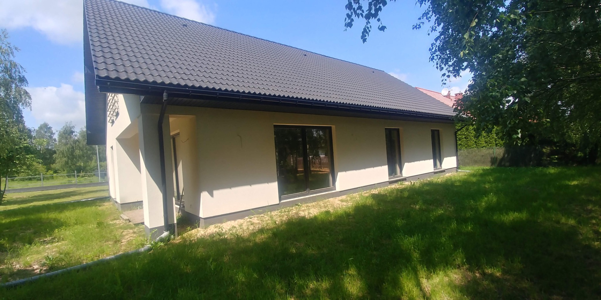 Nowoczesny parterowy dom z szarą dachówką, dużymi oknami i jasną elewacją, otoczony zielonym trawnikiem i drzewami w słoneczny dzień.