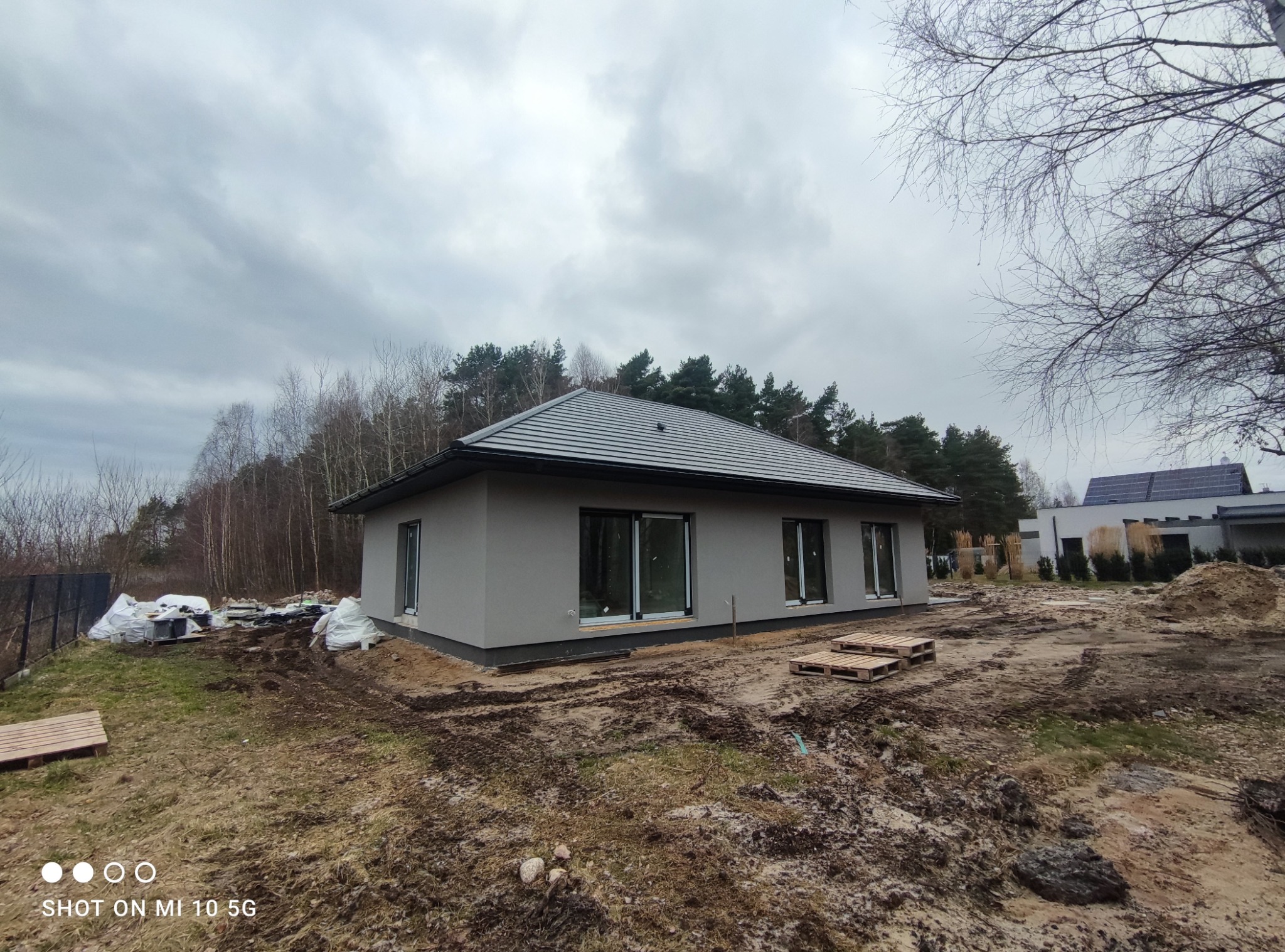 Nowoczesny, parterowy dom z szarą elewacją i grafitową dachówką, w trakcie prac wykończeniowych terenu, widoczne palety i worki z materiałami budowlanymi.