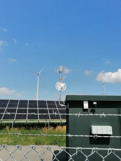 Panel fotowoltaiczny i turbina wiatrowa w tle, widoczne za ogrodzeniem z drutu kolczastego i budynkiem z osprzętem na zewnątrz.