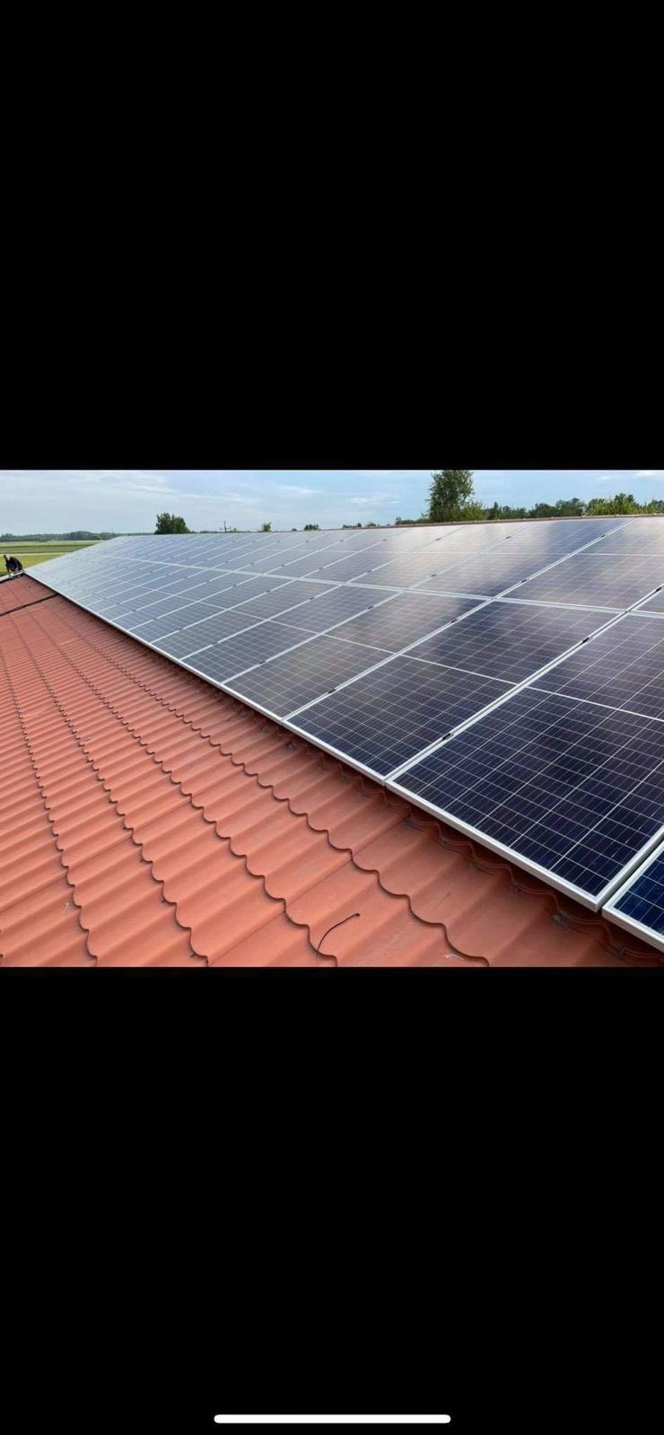 Panele fotowoltaiczne zamontowane na dachu z czerwonej dachówki ceramicznej, widok z boku, w tle zieleń i błękitne niebo.