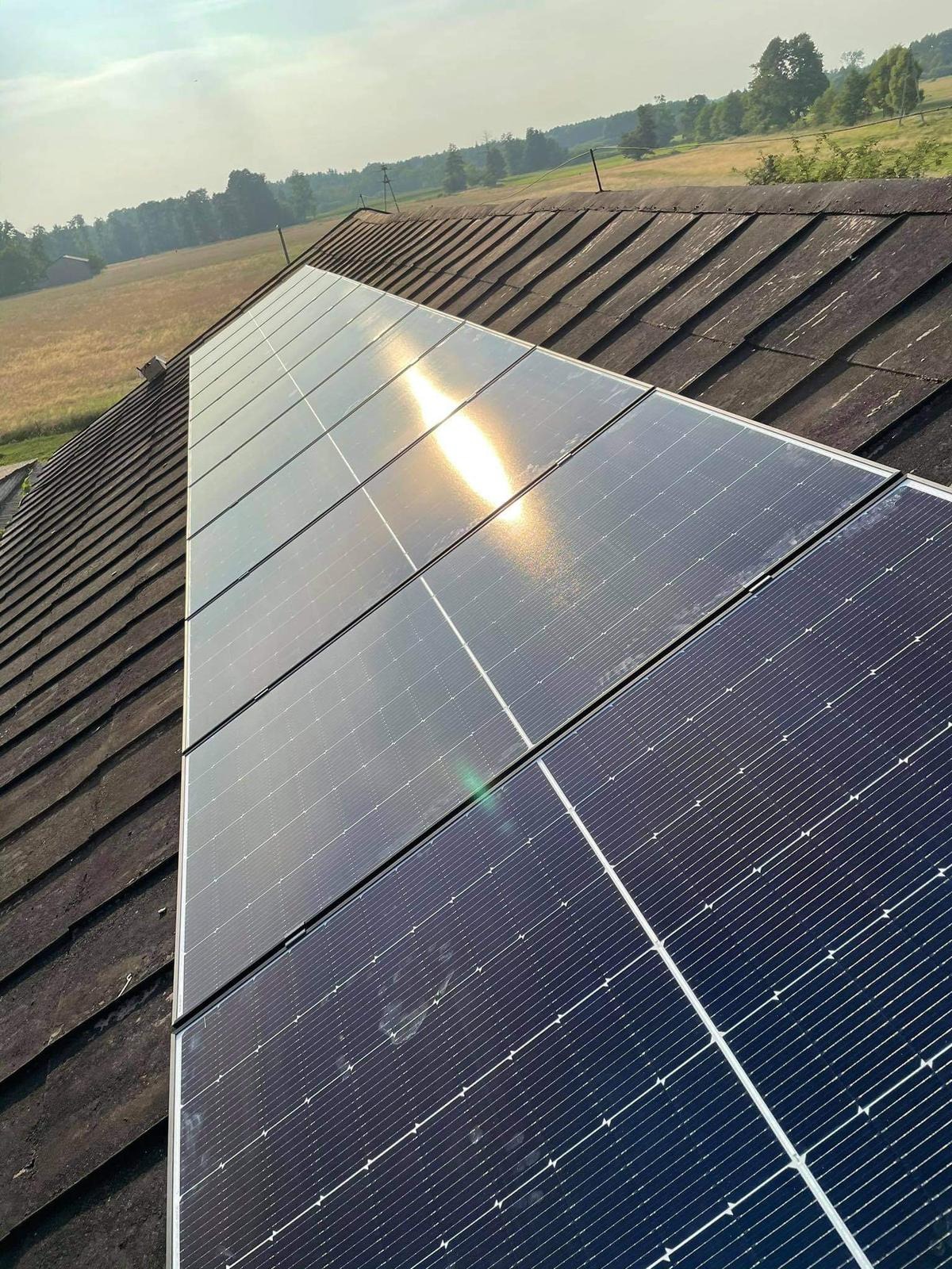 Panele fotowoltaiczne zamontowane na starym, ciemnym dachu z dachówki, widoczne odbicie słońca na panelach, w tle pole i drzewa.