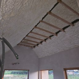 MOSBUD DAWID MOSKAŁA - Poddasze ocieplone pianką PUR z widocznymi drewnianymi belkami, szary wąż do natrysku pianki w rogu, okna z widokiem na zieleń.