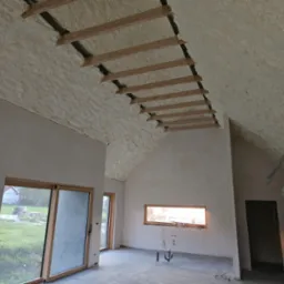 Poddasze domu jednorodzinnego po natrysku izolacji, widoczne drewniane elementy konstrukcyjne dachu wystające spod warstwy izolacyjnej, okno z widokiem na ogród i przewody wentylacyjne.