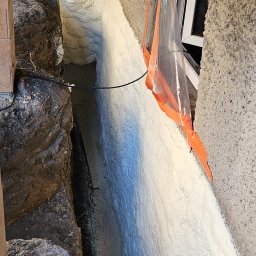 MOSBUD DAWID MOSKAŁA - Świeżo nałożona, jasna pianka izolacyjna wypełnia szczelinę fundamentu, z widocznym zabezpieczeniem okna folią malarską z pomarańczową taśmą.