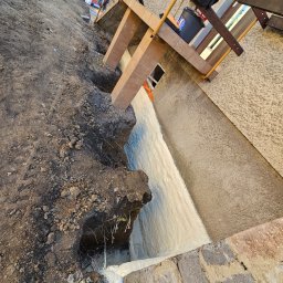 MOSBUD DAWID MOSKAŁA - Wykonana izolacja fundamentów pianą PUR w wykopie, widoczne ślady ziemi i korzenie, zabezpieczenie budynku rusztowaniem z desek.