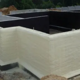 Fundamenty budynku zabezpieczone czarną hydroizolacją i pokryte warstwą jasnej pianki izolacyjnej, widoczne wystające pręty zbrojeniowe i teren budowy w tle.