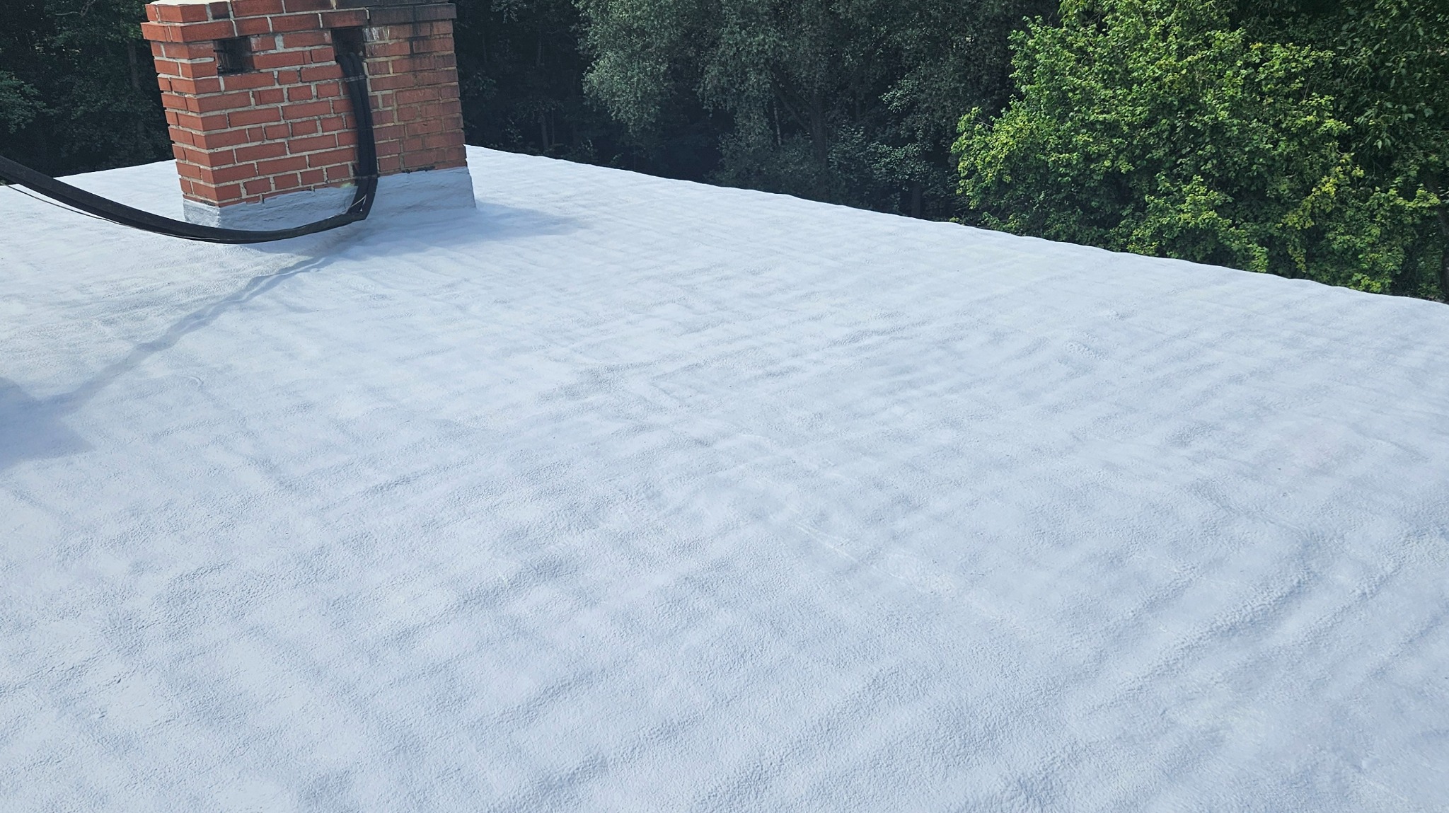 Szary dach pokryty jednolitą warstwą izolacyjną z widocznym kominem z cegły i fragmentami zieleni w tle.