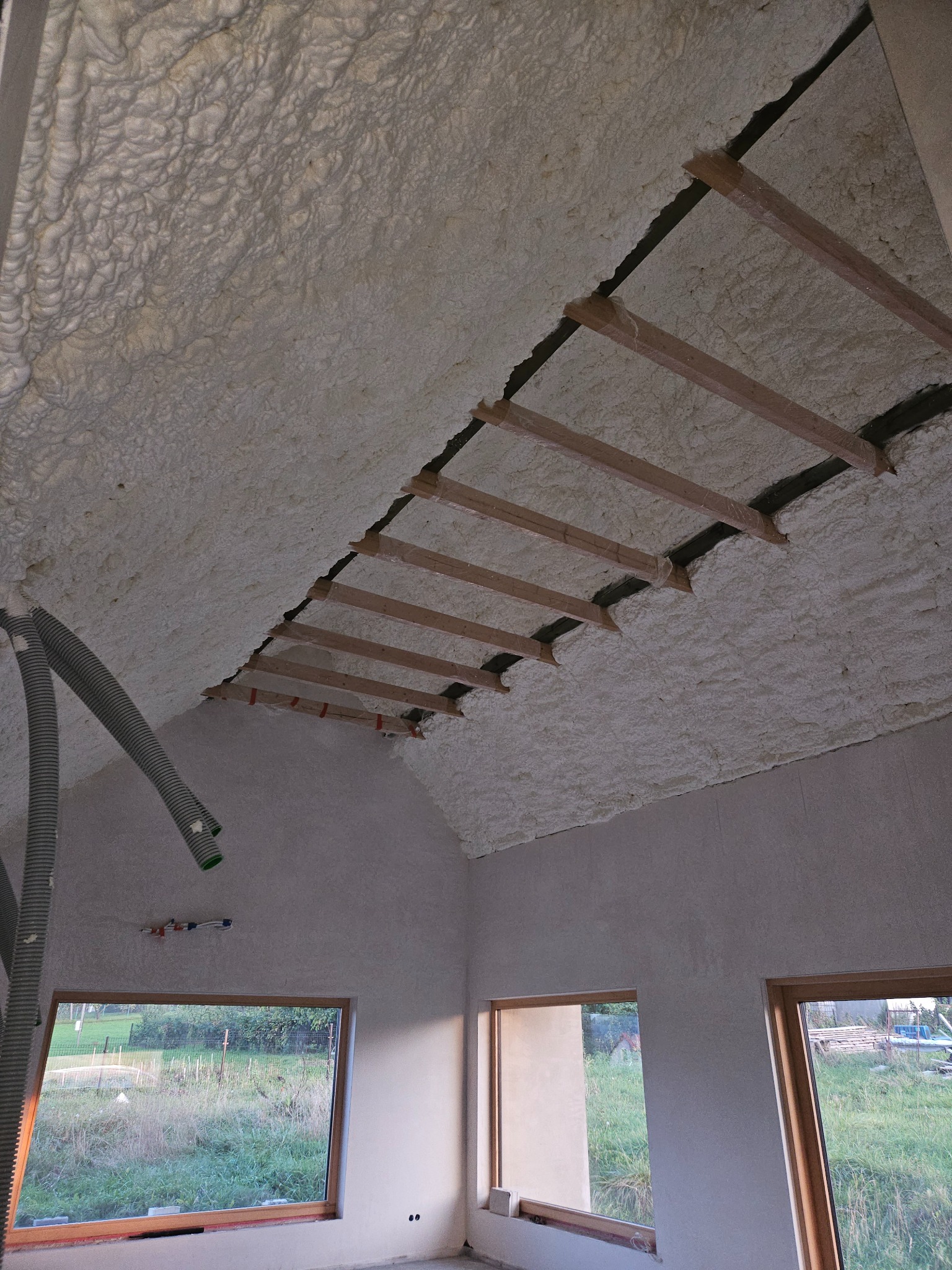 Poddasze ocieplone pianką PUR z widocznymi drewnianymi belkami, szary wąż do natrysku pianki w rogu, okna z widokiem na zieleń.