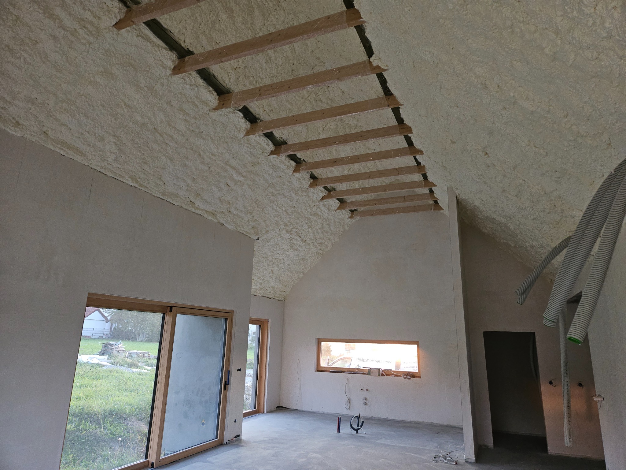 Poddasze domu jednorodzinnego po natrysku izolacji, widoczne drewniane elementy konstrukcyjne dachu wystające spod warstwy izolacyjnej, okno z widokiem na ogród i przewody wentylacyjne.