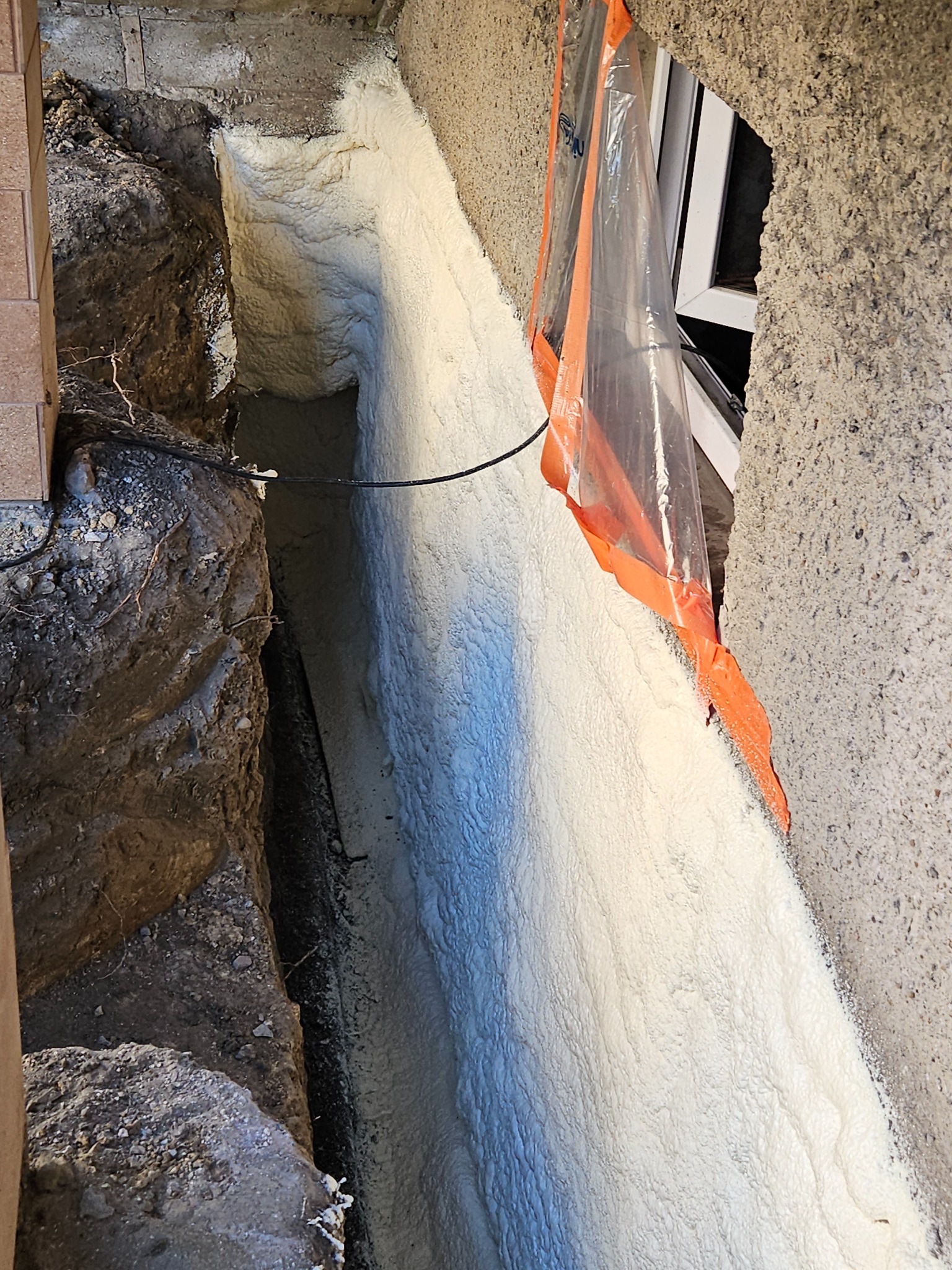 Świeżo nałożona, jasna pianka izolacyjna wypełnia szczelinę fundamentu, z widocznym zabezpieczeniem okna folią malarską z pomarańczową taśmą.
