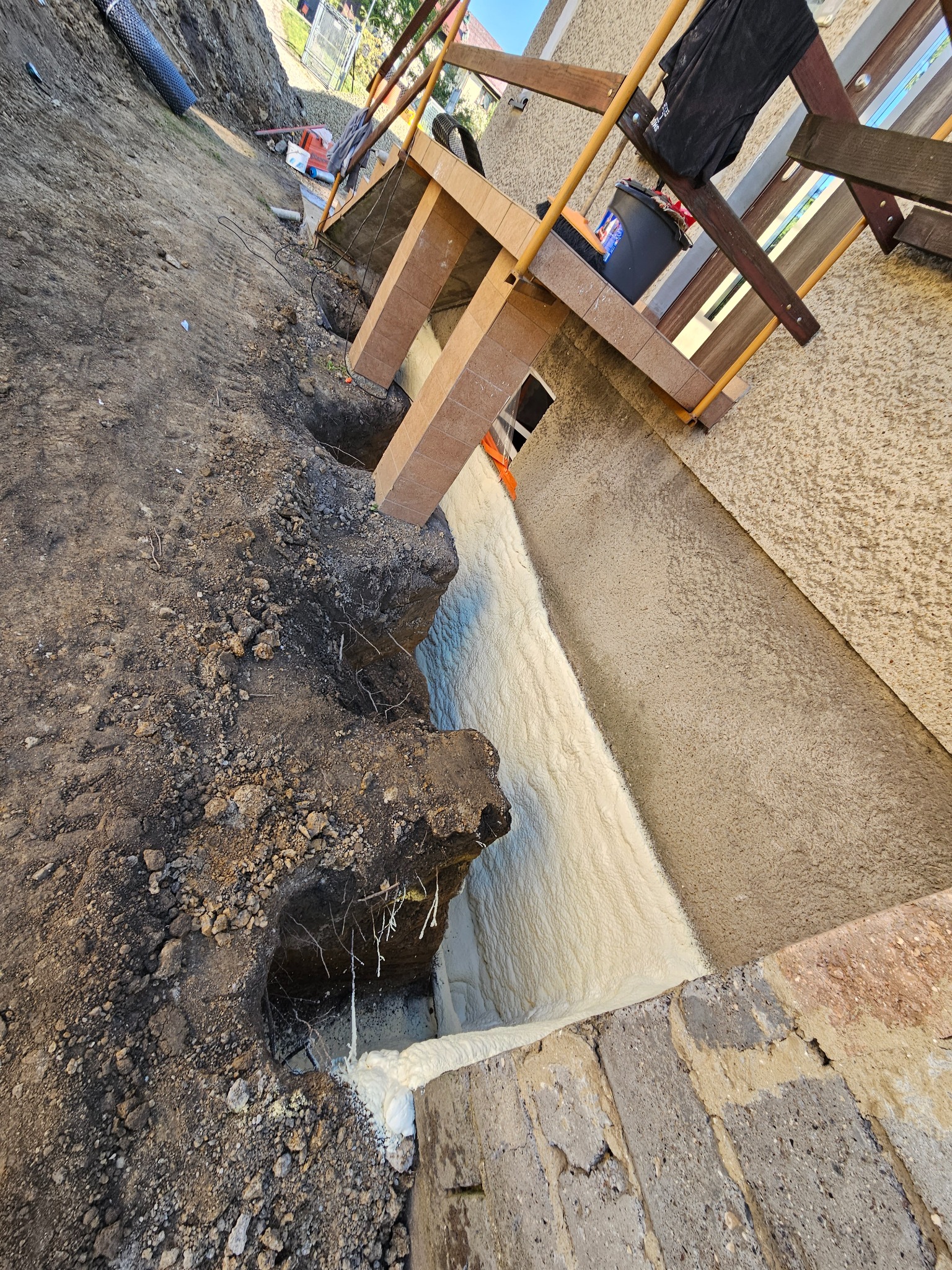 Wykonana izolacja fundamentów pianą PUR w wykopie, widoczne ślady ziemi i korzenie, zabezpieczenie budynku rusztowaniem z desek.